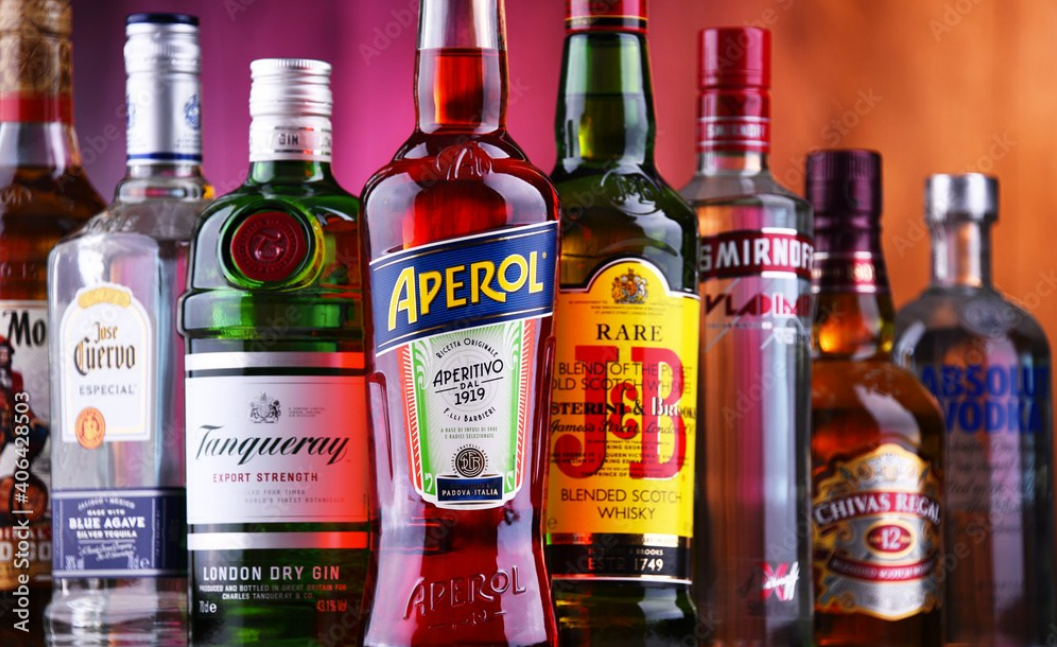 Assorted liquor bottles lined up on a bar shelf, including a red Aperol bottle in the center.