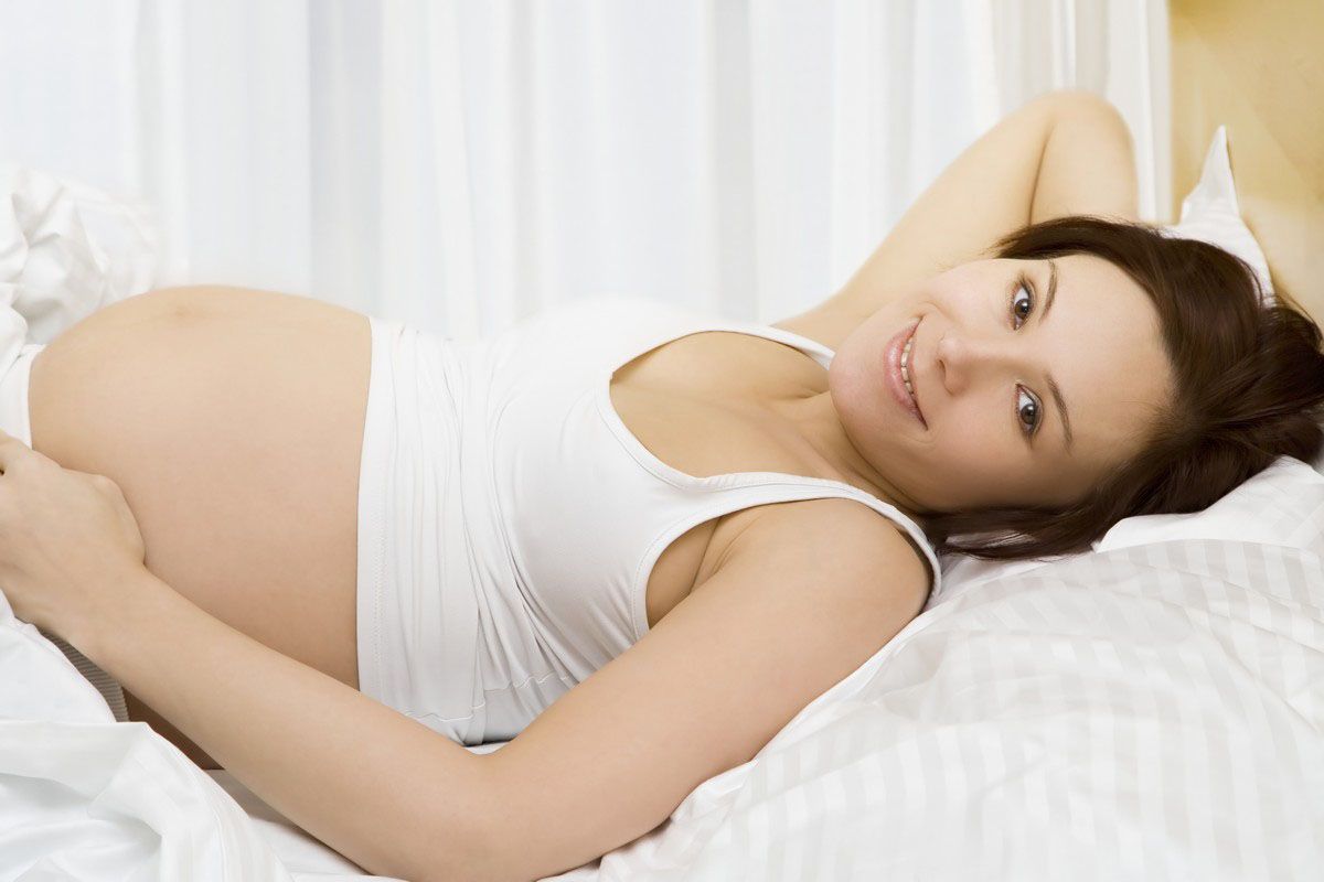 Pregnant person lying in bed, smiling, holding belly. Wearing white tank top.
