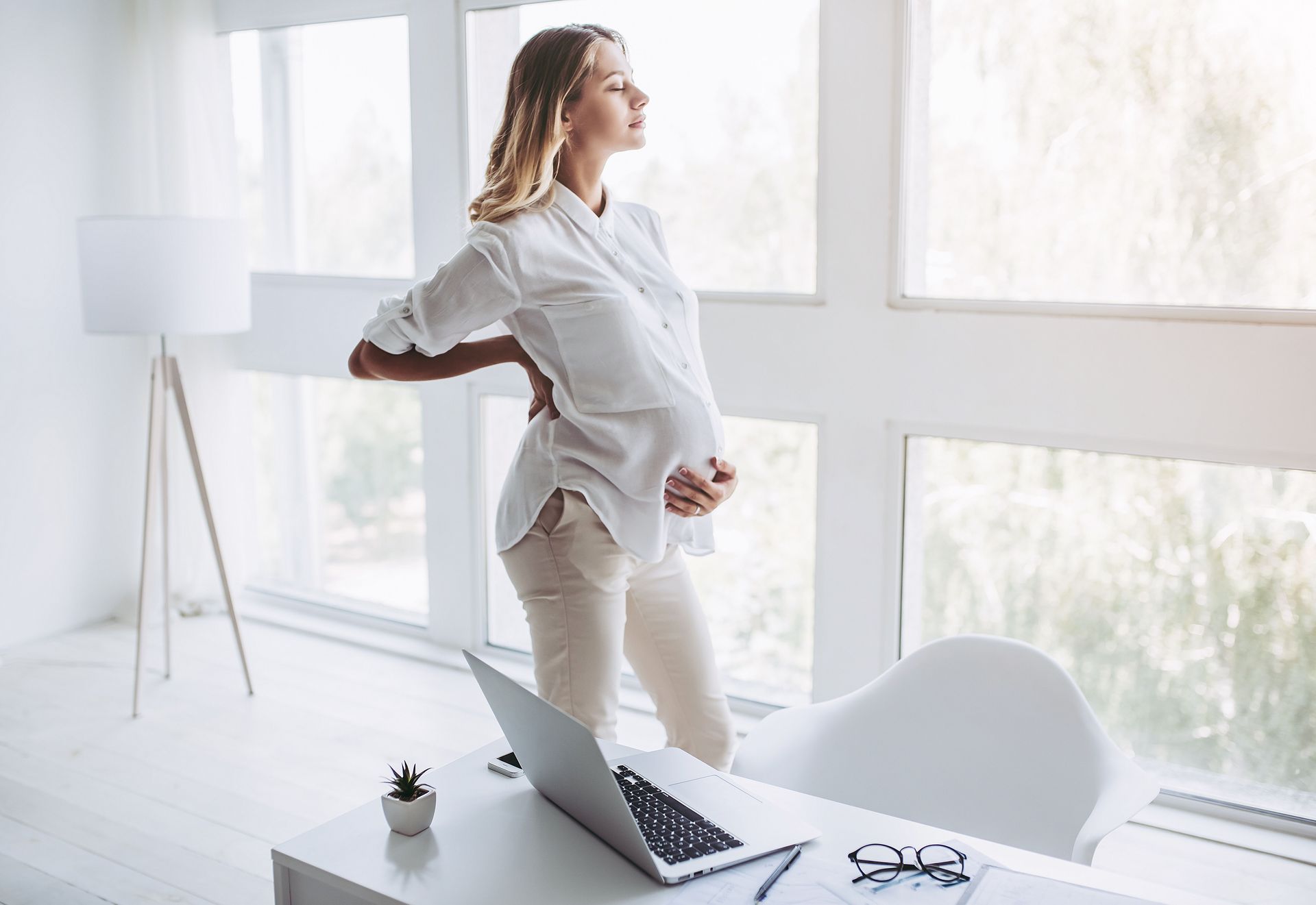 Pregnant person holding back, standing by window, looking away from desk with laptop.