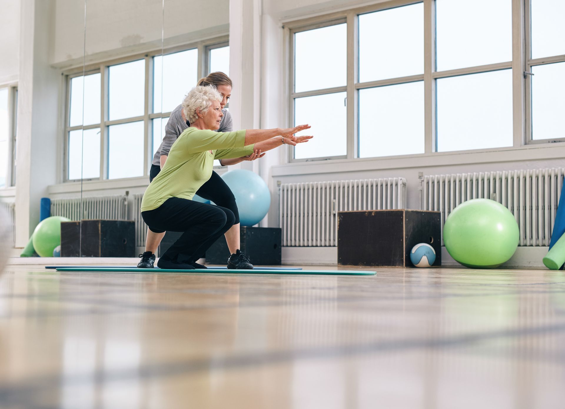 An elderly woman is squatting on a mat in a gym with a personal trainer.