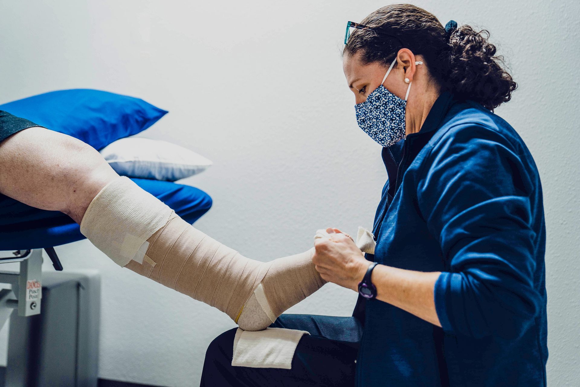 A nurse is bandaging a patient 's leg while wearing a mask.
