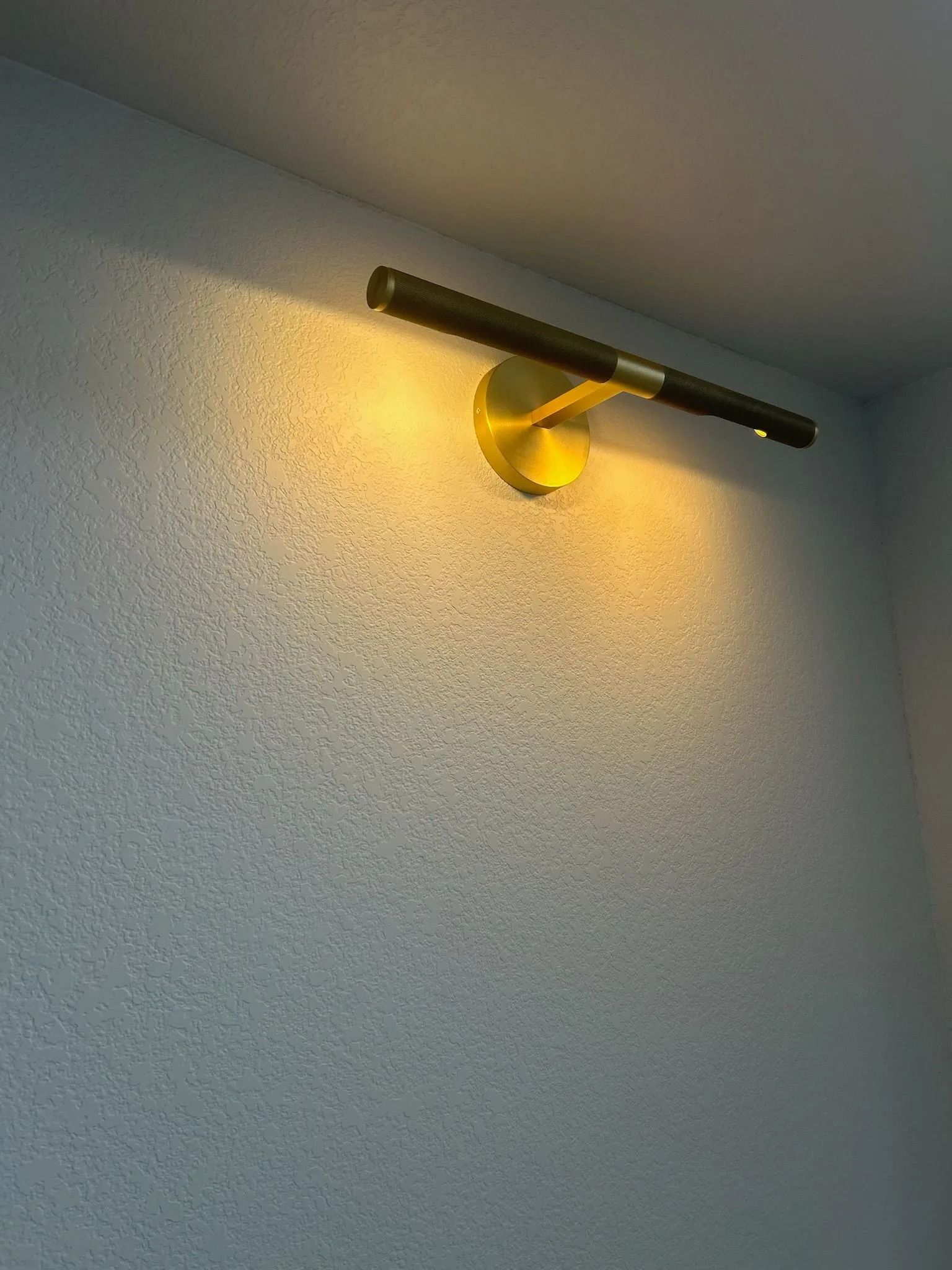 Brass wall lamp with a long, black light bar on a textured, light gray wall.