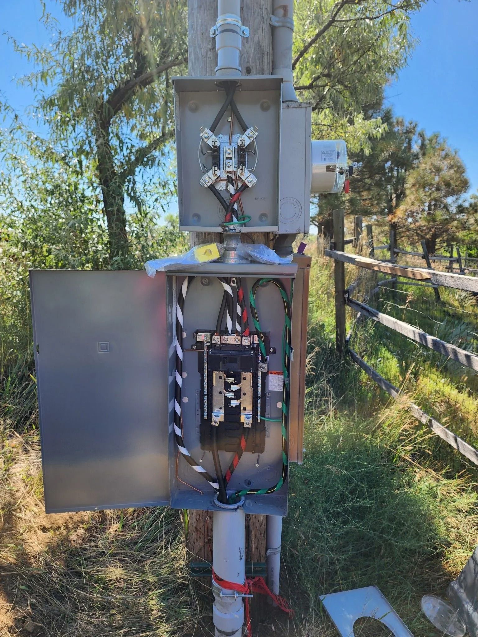 Opened electrical box on a pole with wires, breakers, and conduits, outdoors in a grassy area.