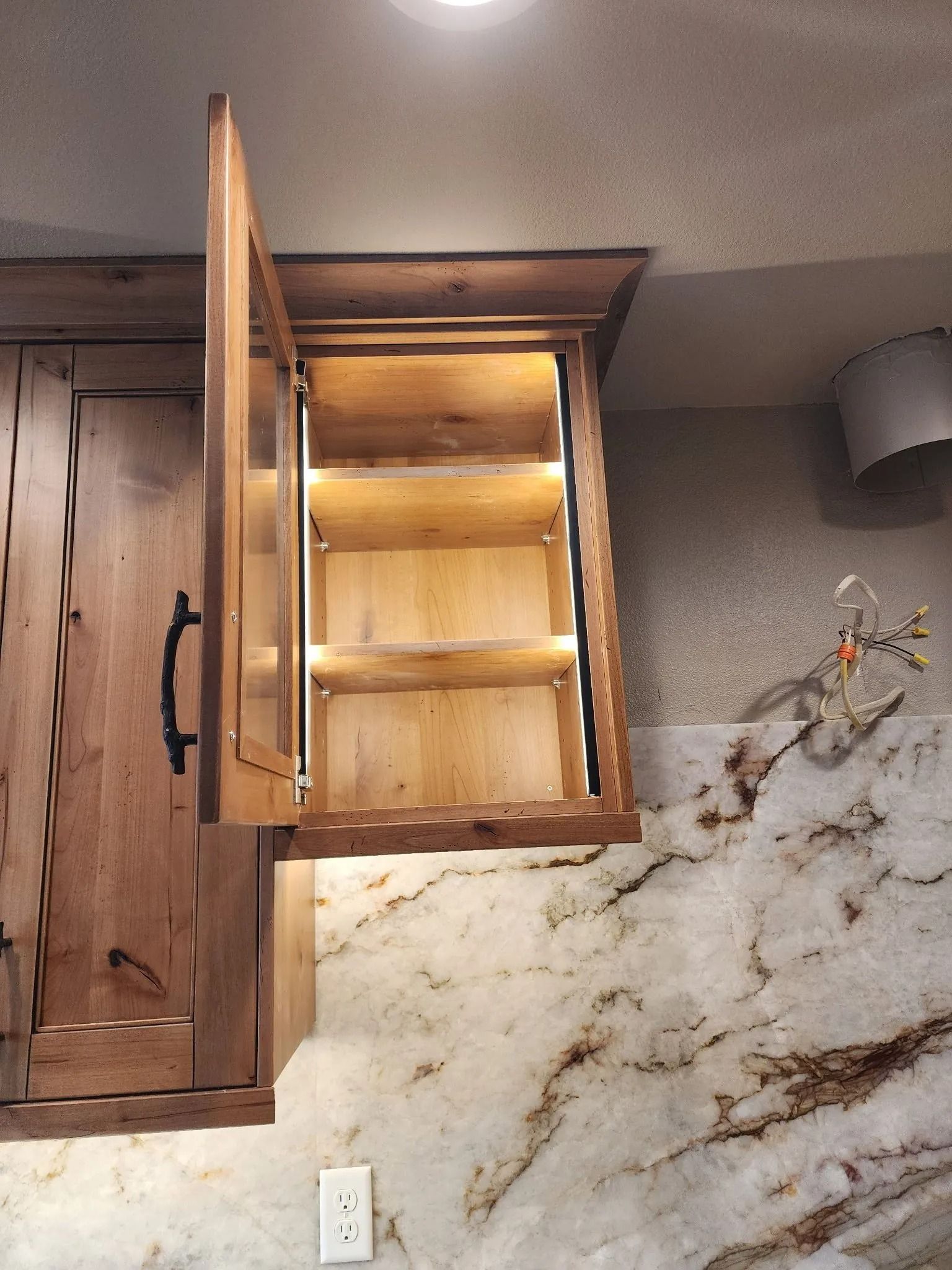 Wooden cabinet with glass door open, revealing lit shelves above a granite countertop.