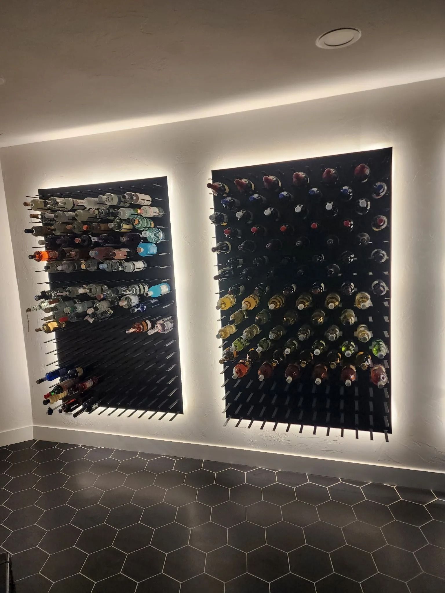 Two wall-mounted wine racks with bottles, lit by strip lighting, against a white wall. Black tile floor.