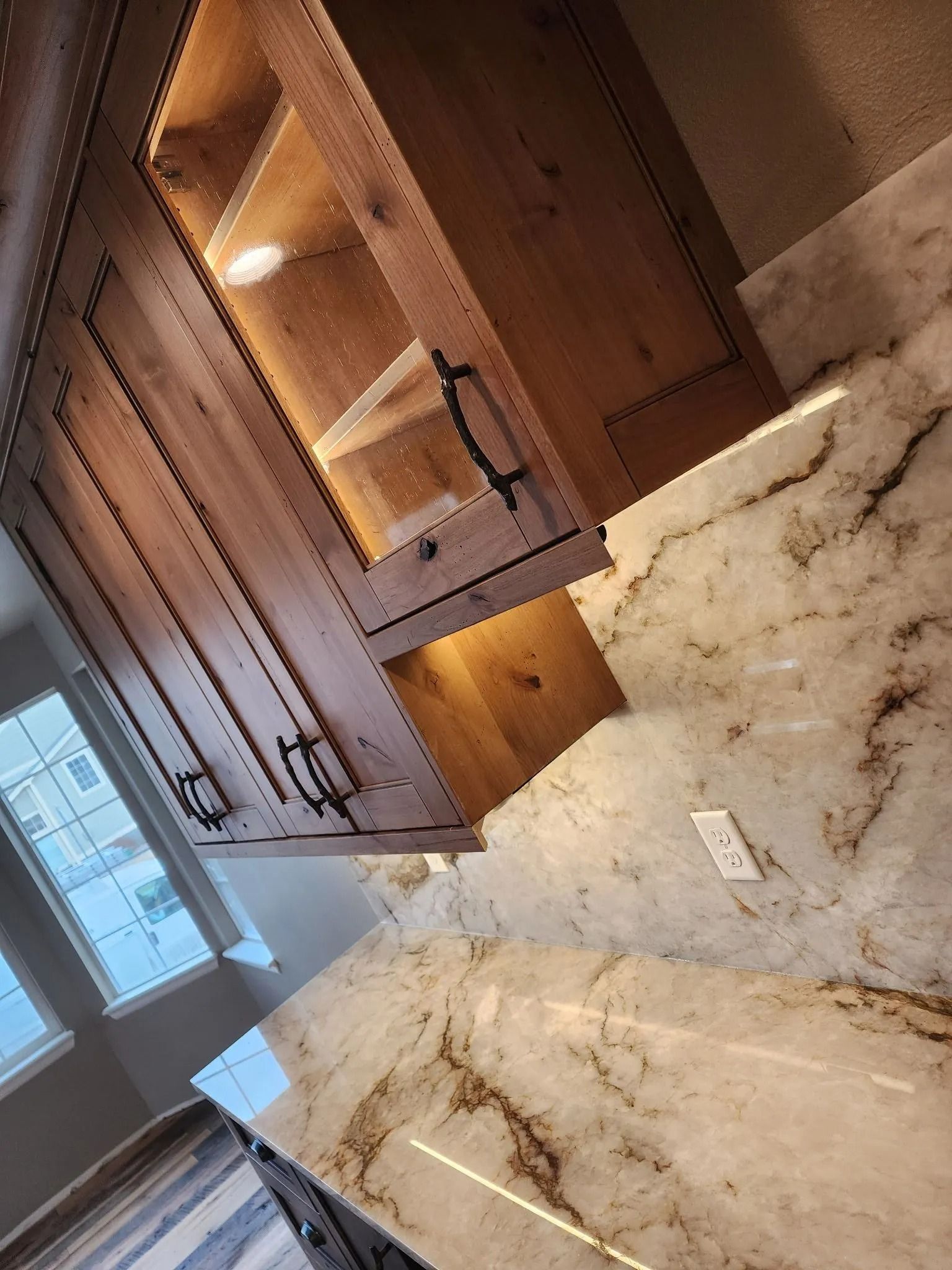 Wooden kitchen cabinet with glass front, lit interior, over marble countertop.