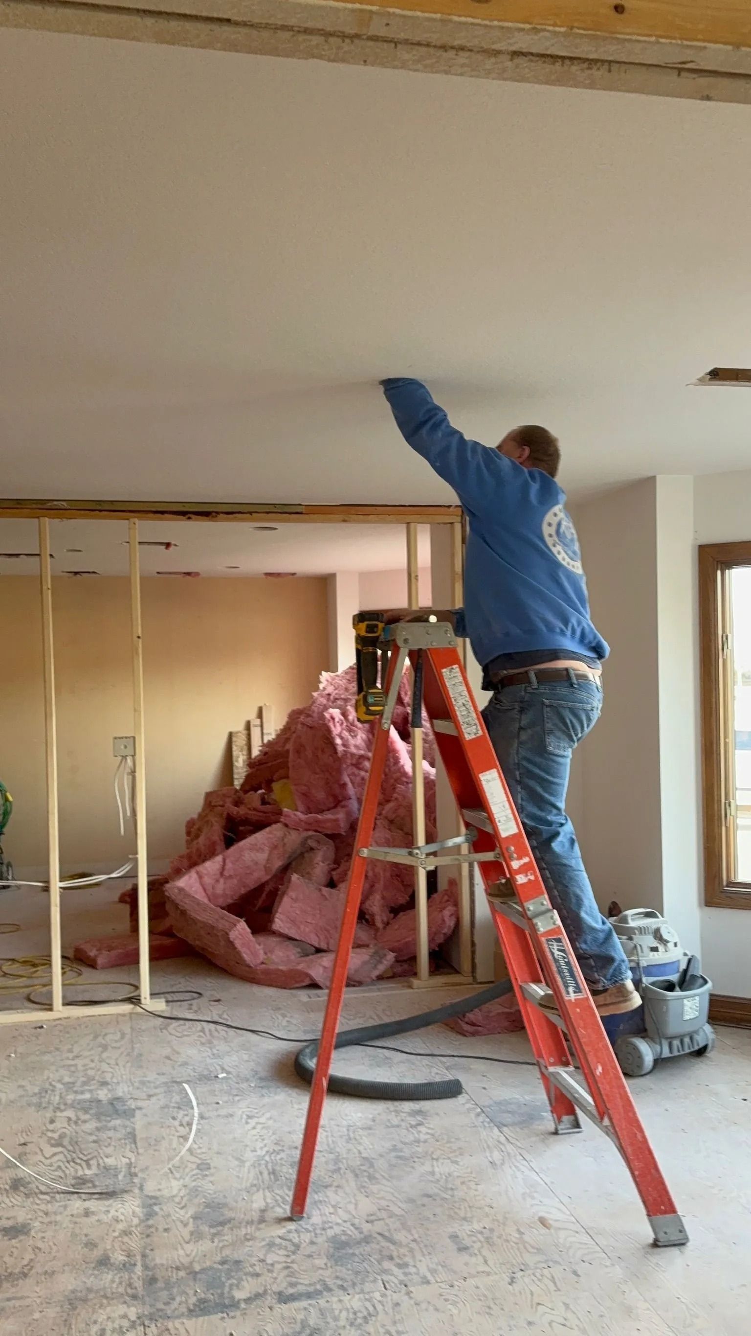 Person on a ladder working on a ceiling during a renovation. Interior setting with exposed wall studs and insulation.