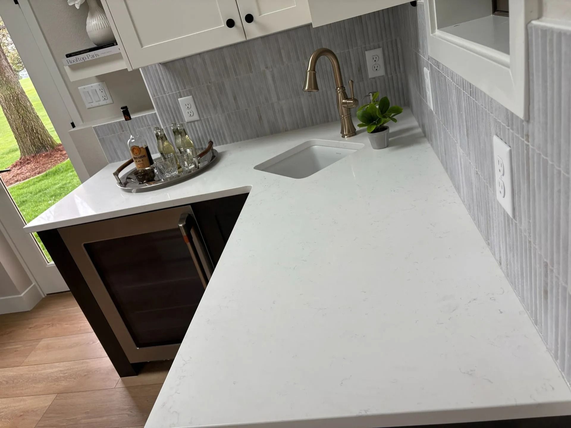 White countertop with sink, gold faucet, and textured gray backsplash. Built-in wine cooler below.