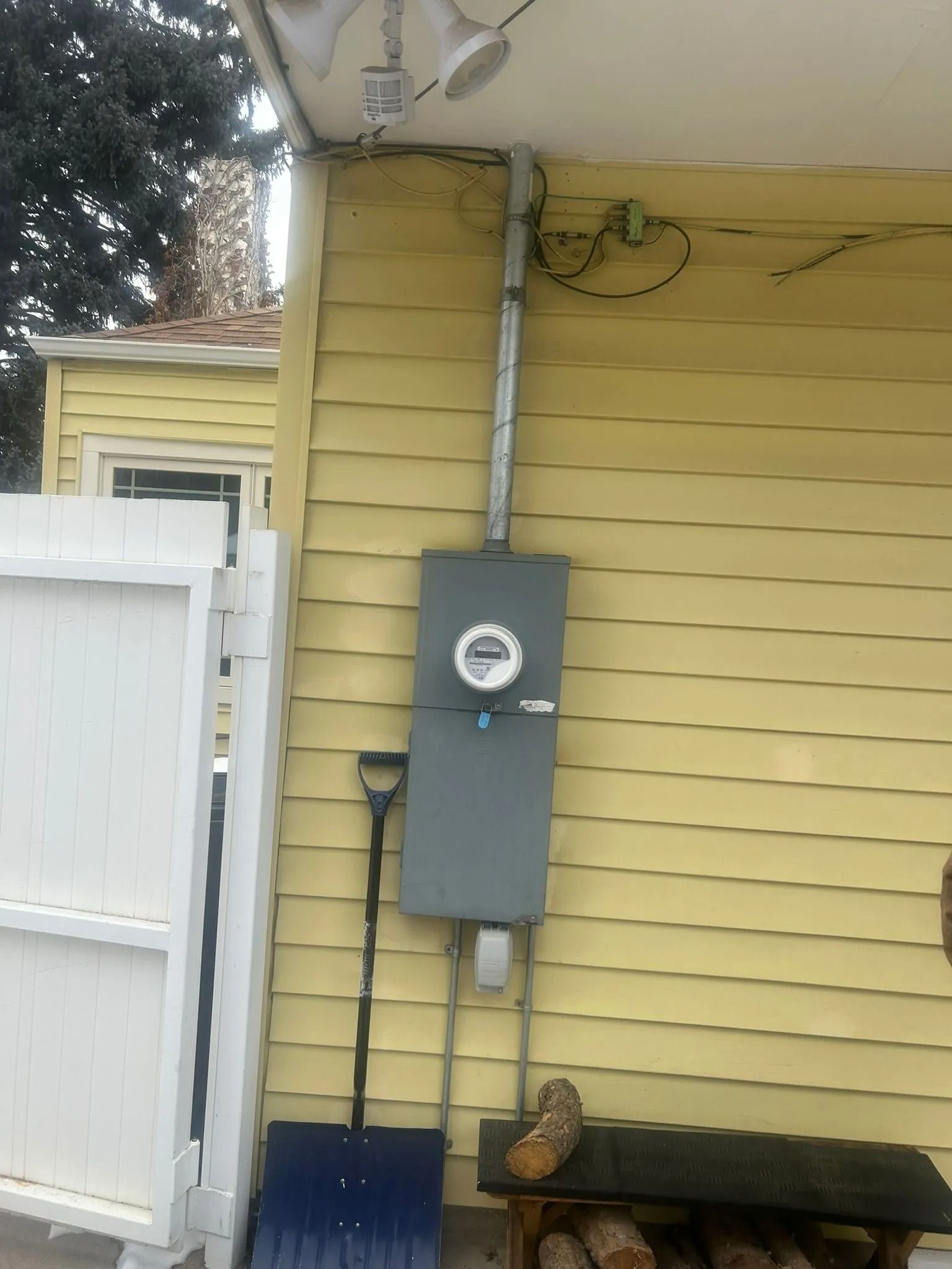 Exterior electrical meter box on a yellow house wall with a shovel, wood, and a white fence.