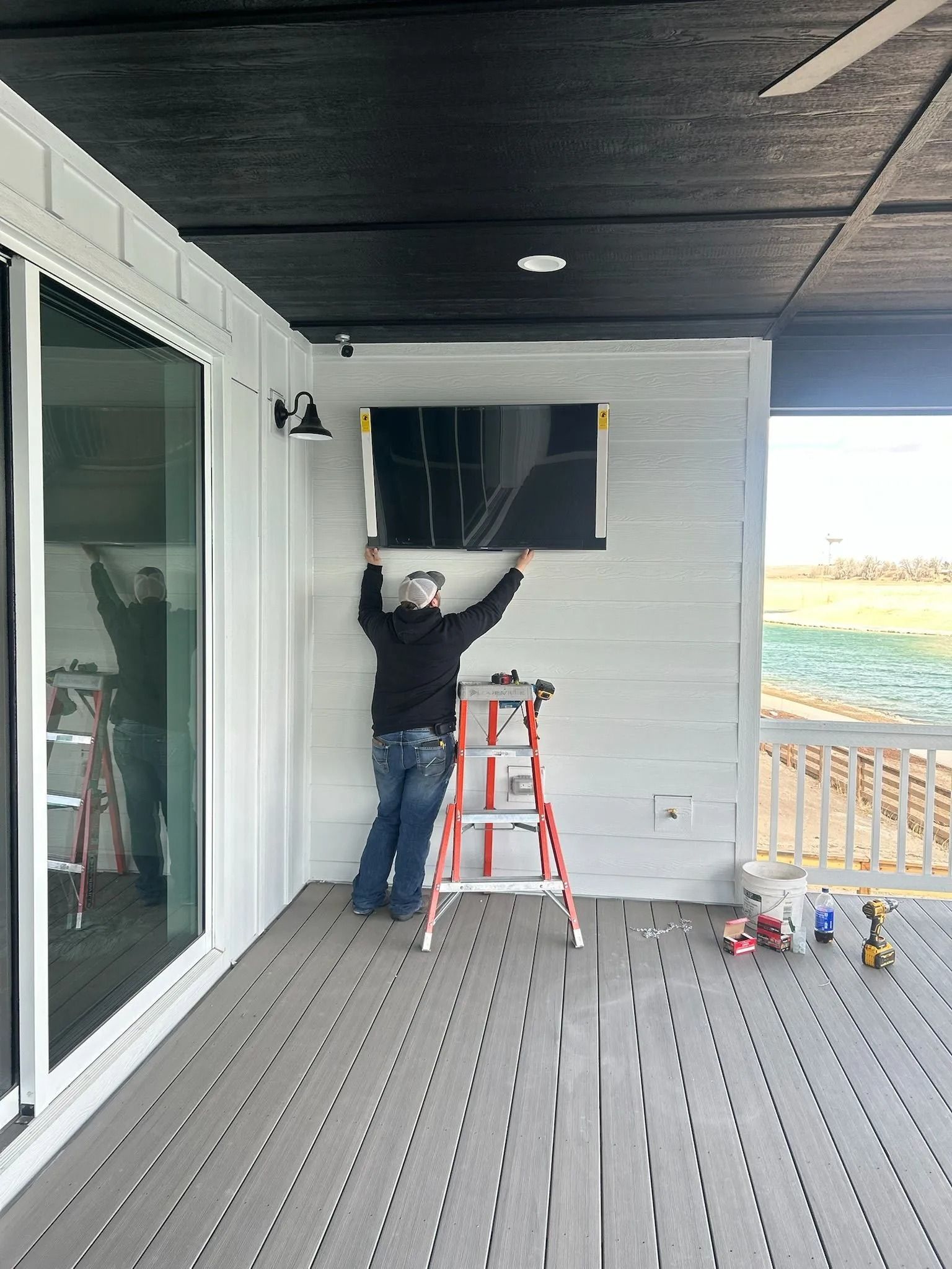 Person on ladder mounting a TV on a white-paneled exterior wall, outdoor deck, gray composite decking, black ceiling.