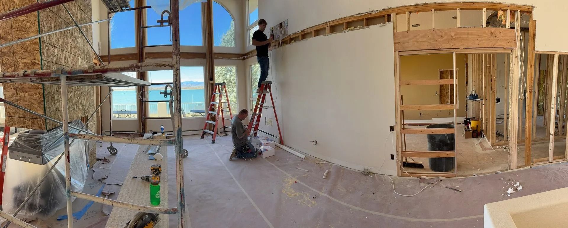 Construction workers renovating a room with large windows overlooking water. One worker on a ladder, another on the floor.