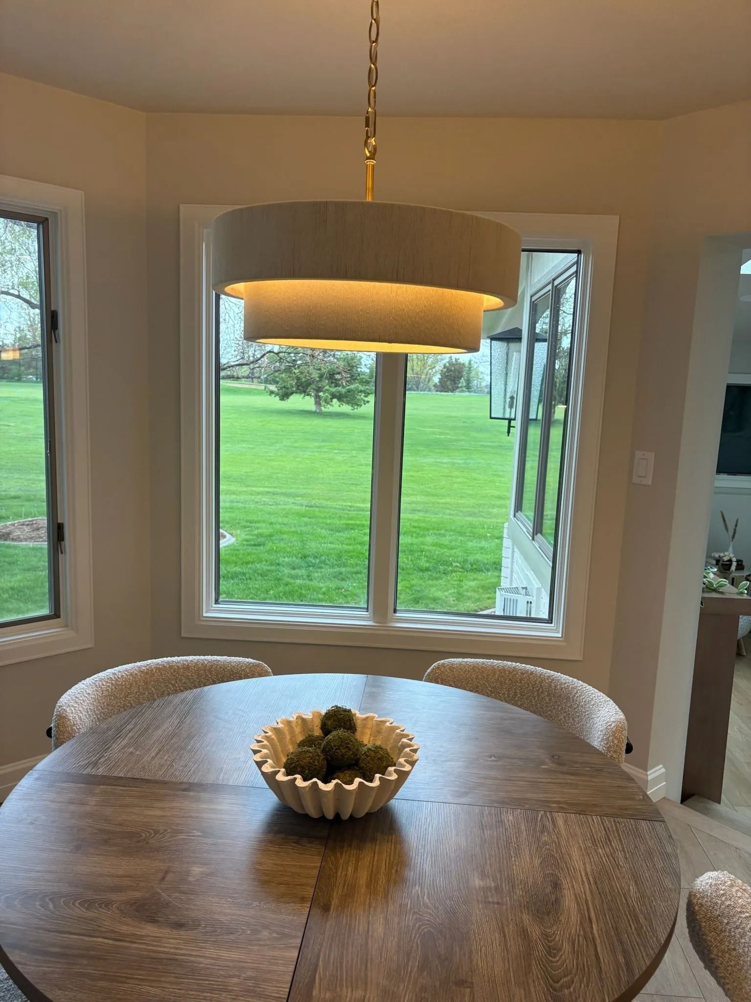 A round wooden dining table with a decorative bowl beneath a tiered beige pendant light by a large window overlooking a lawn.