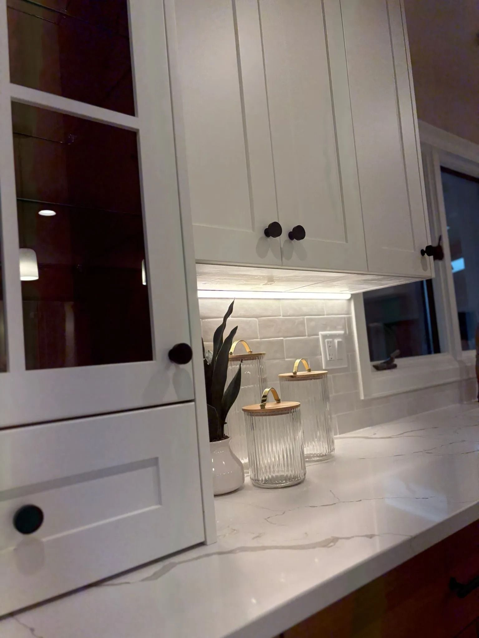 White kitchen cabinets with black knobs, under-cabinet lighting, and glass jars on a marble countertop.