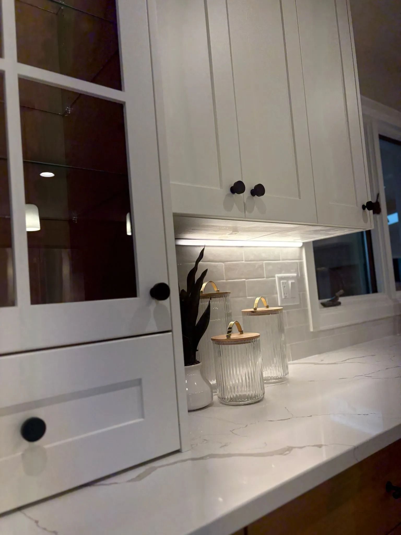 White kitchen cabinets with black knobs, under cabinet lighting, and glass jars on a countertop.