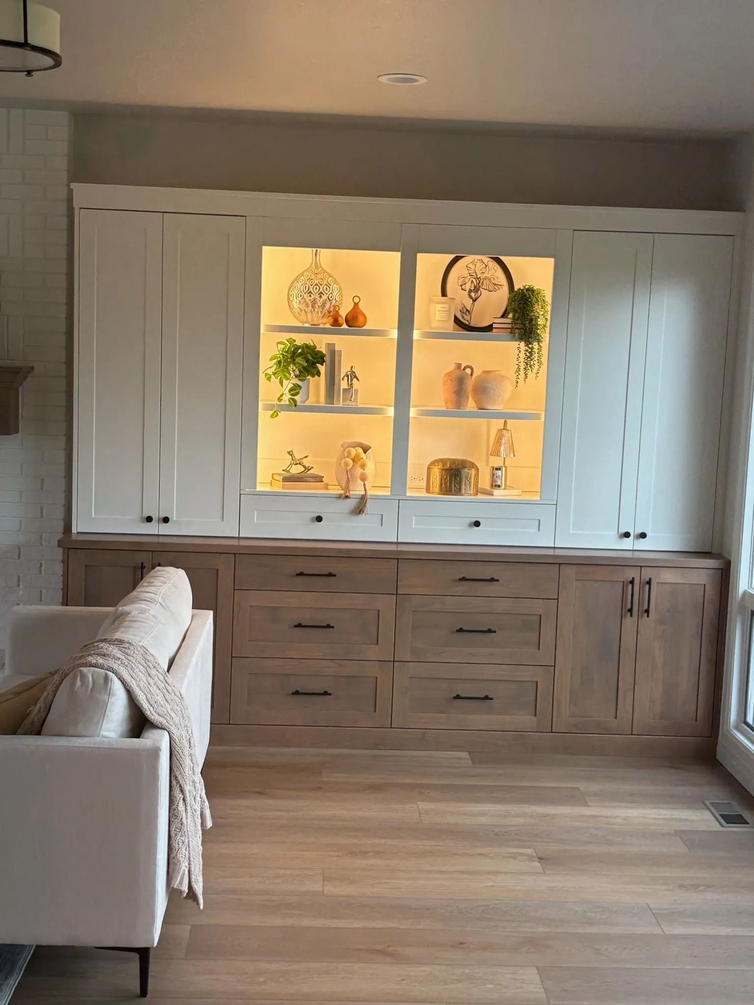 Built-in cabinet with white upper cabinets, lit shelves, and light wood drawers and lower cabinets; sofa on the left.