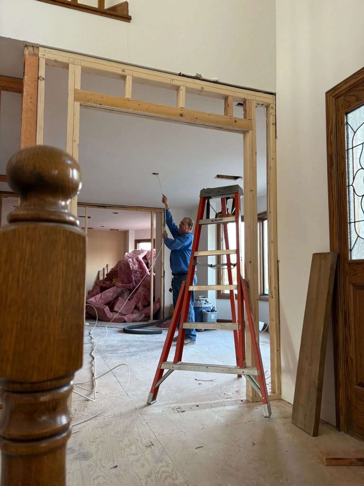 Person on a ladder, working on a door frame in a room under construction.