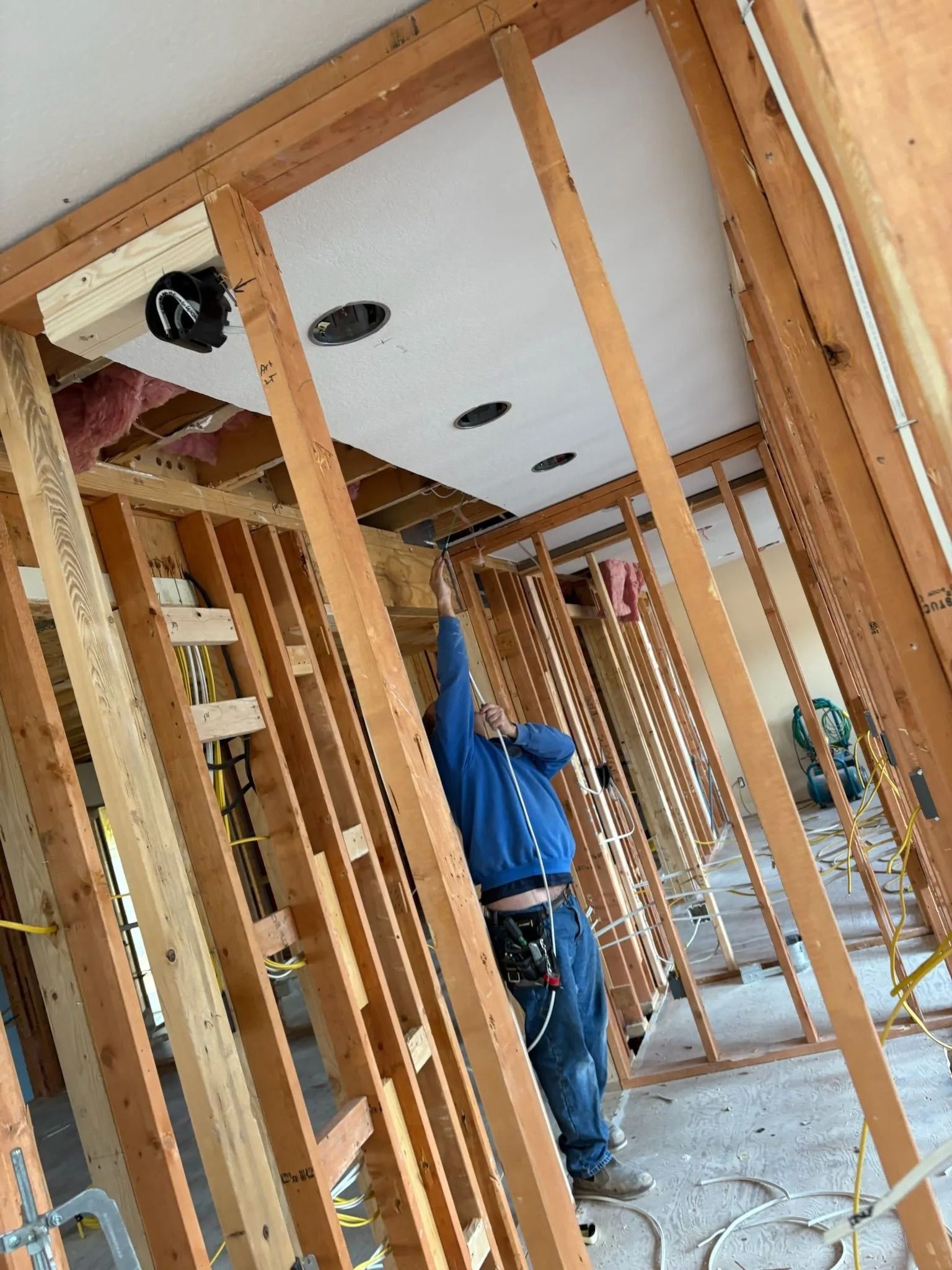Construction worker reaching up at ceiling with recessed lights, wooden framing visible.