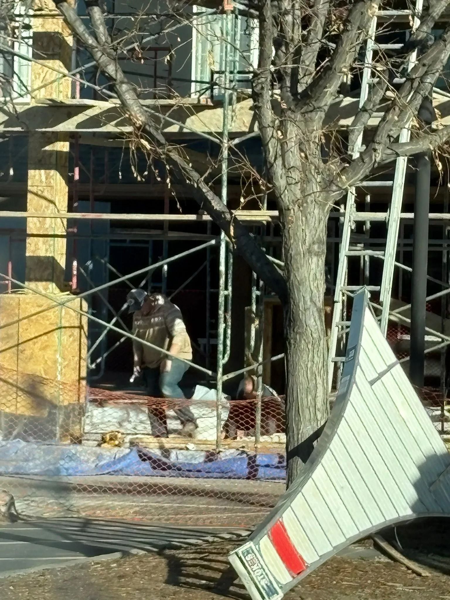 Construction site with scaffolding and workers near a building with exposed wood. A tree and road sign are in the foreground.