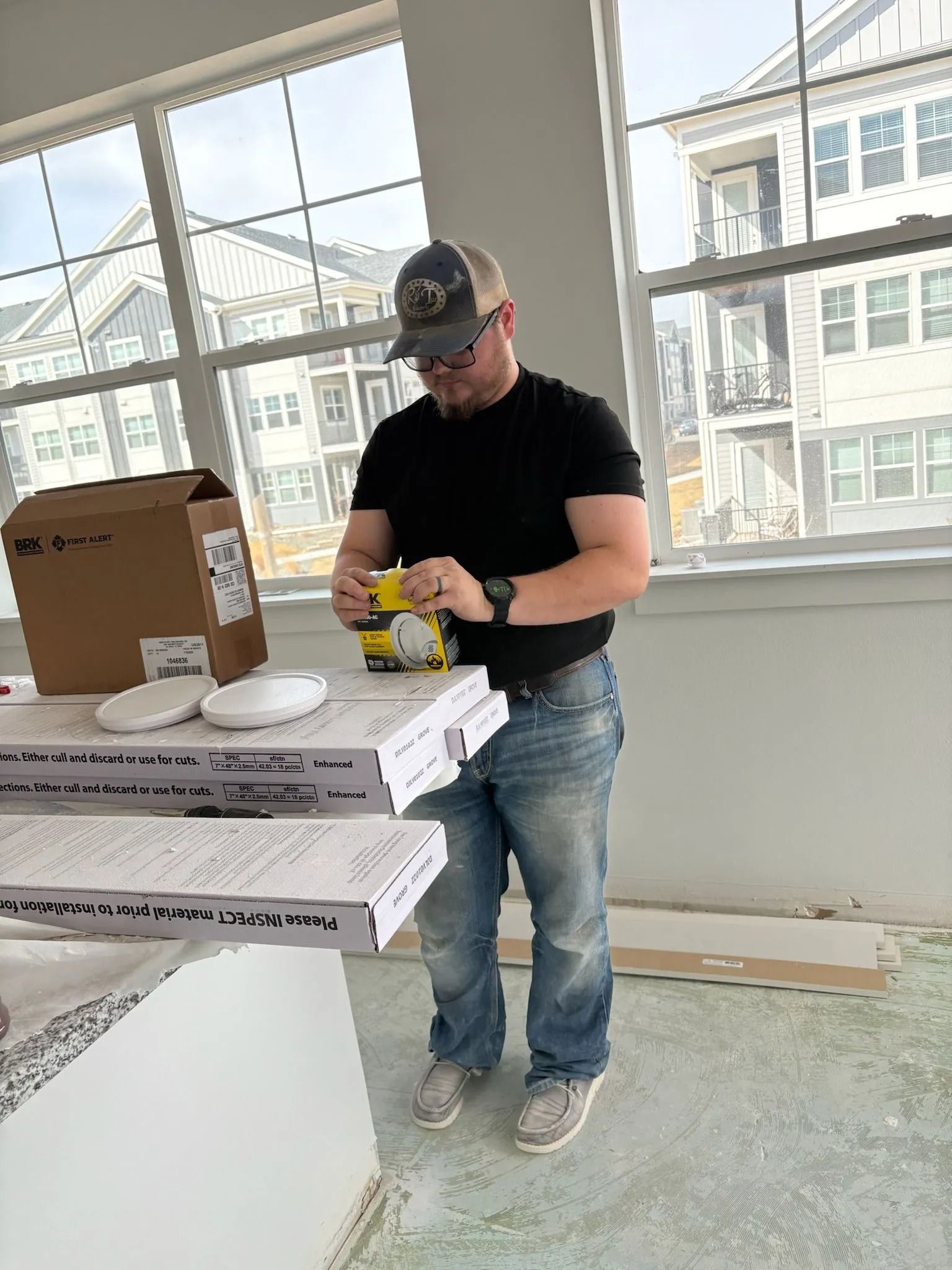Man in jeans and hat using tool near stacks of papers by window in building.