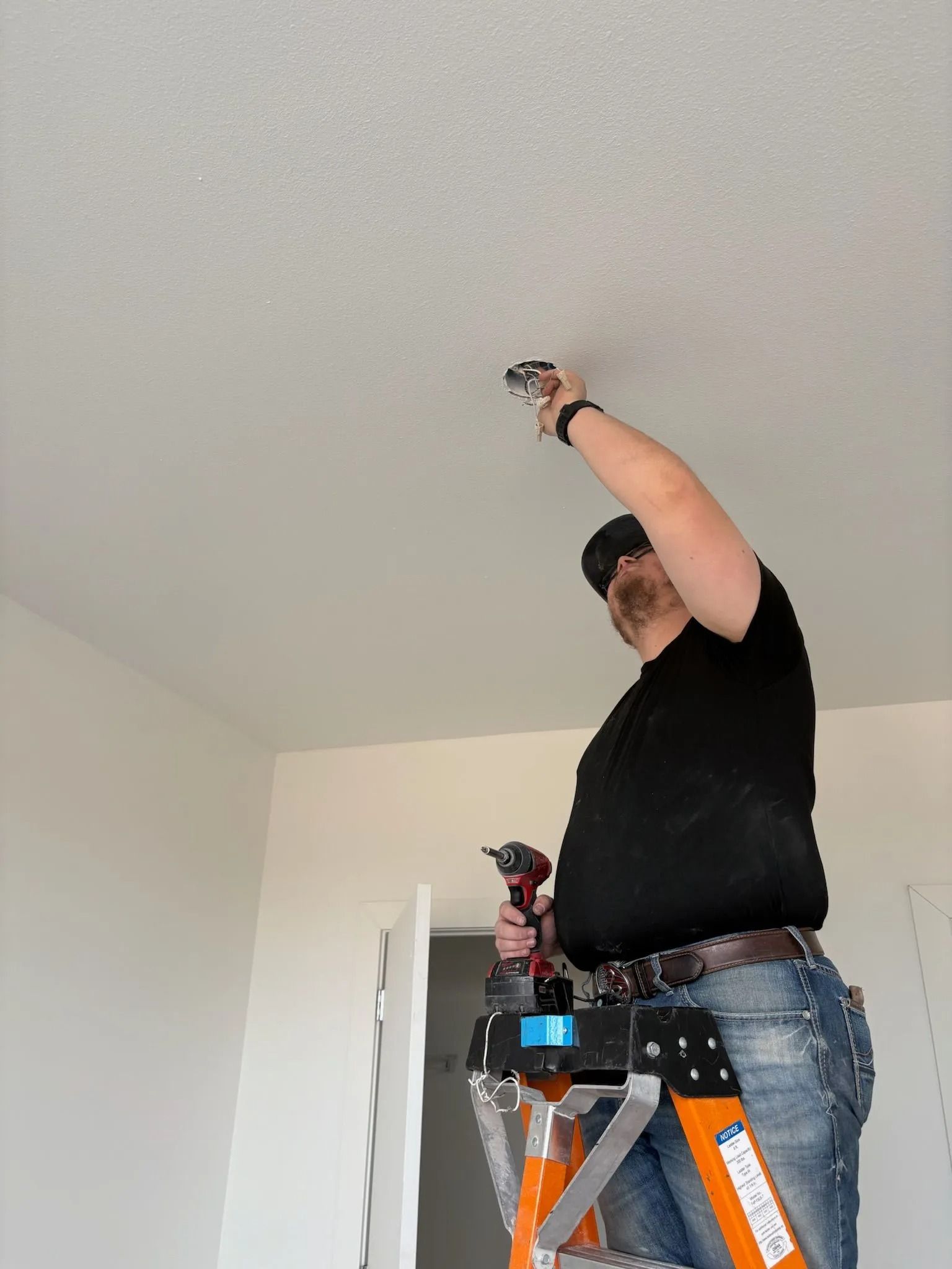 Man on a ladder installing a ceiling fixture, using a drill. He wears a black shirt and jeans.