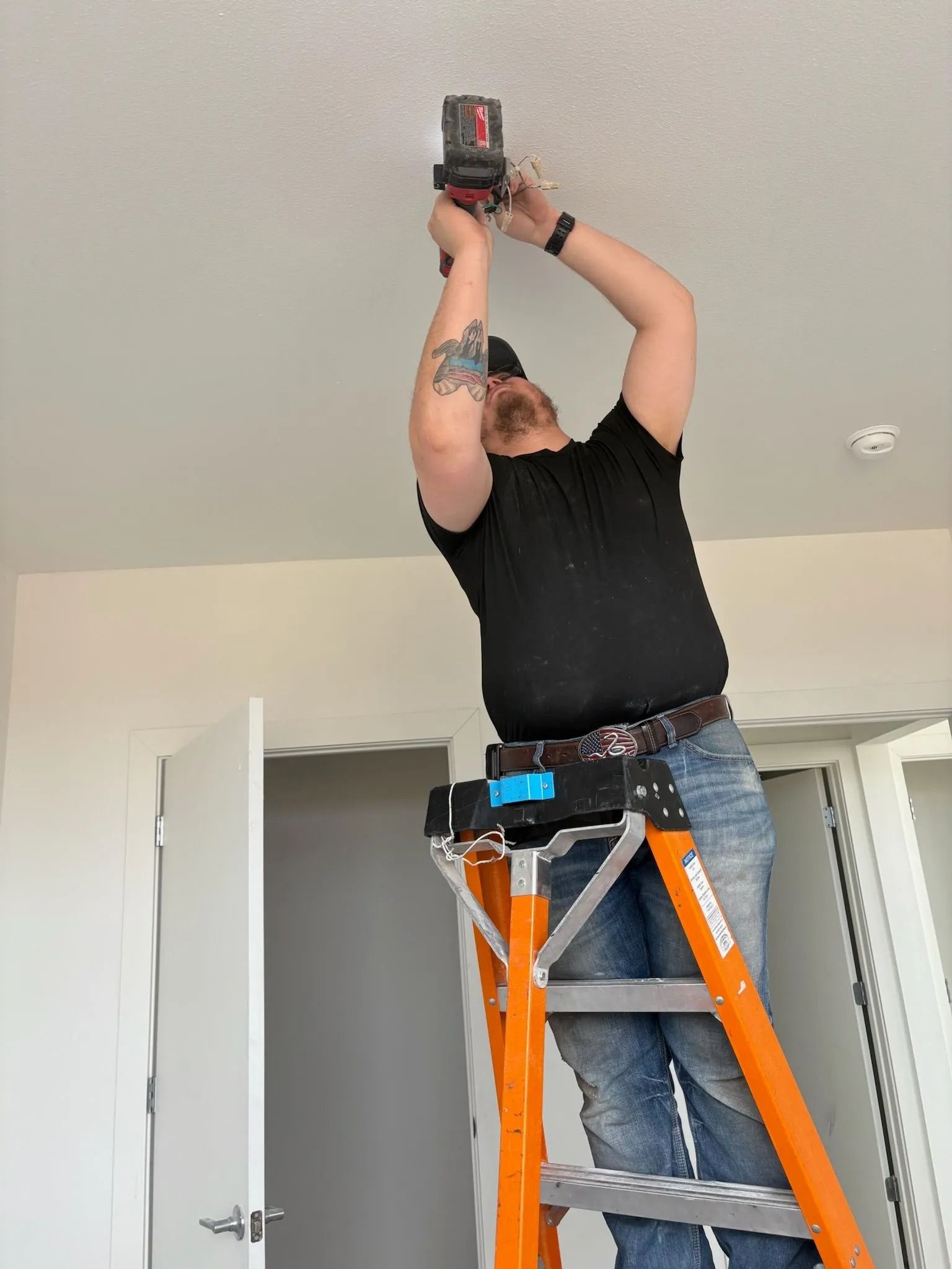 Person on a ladder installing something on a white ceiling. They are holding a tool up with both hands.