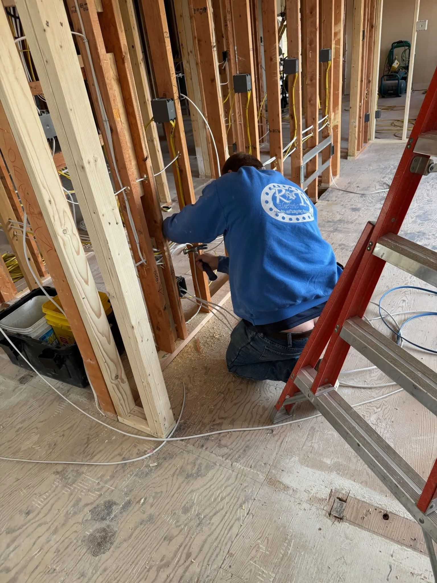Man in blue sweatshirt wiring electrical boxes in a wood-framed wall, next to a red ladder.