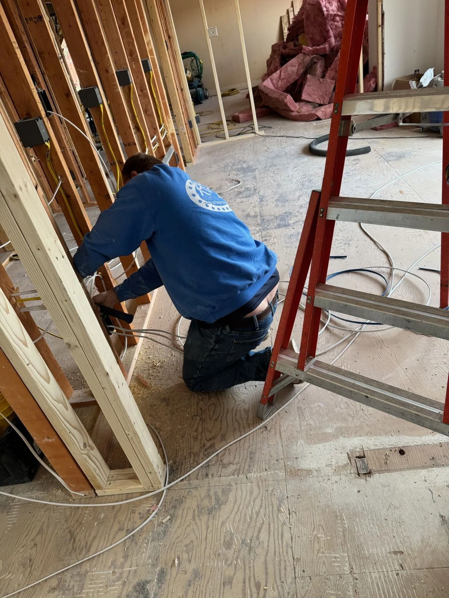Construction worker kneels, wiring in a framed wall with a ladder nearby.