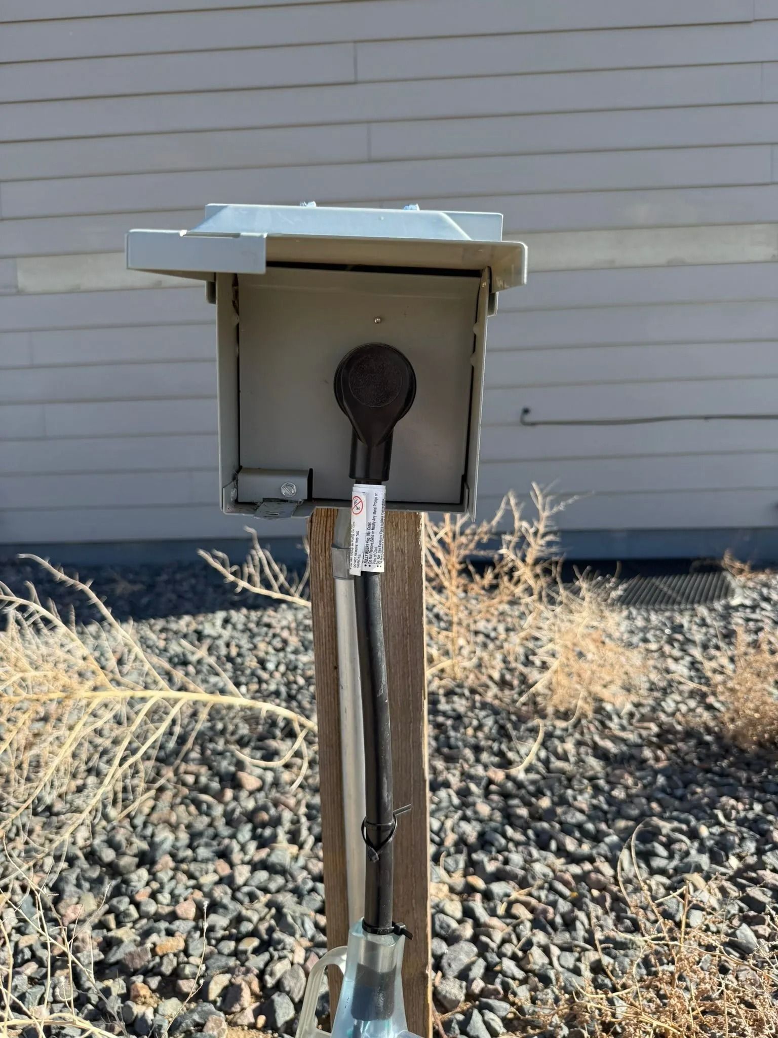 Gray electrical box on a wooden post with a black cable, set outdoors in front of a gray building and rocky ground.