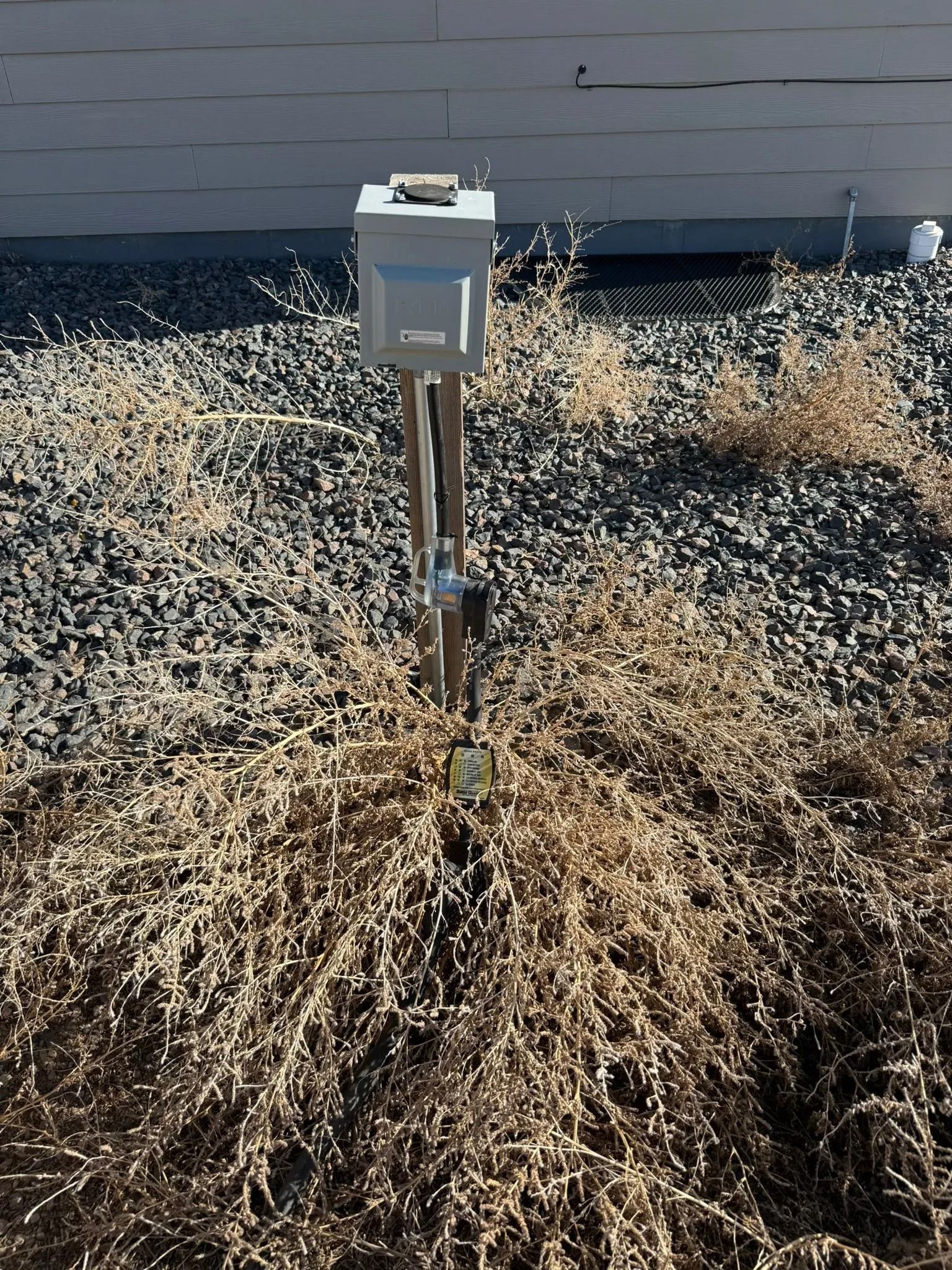 Electrical box on a metal pole surrounded by dry, brown vegetation and gravel.