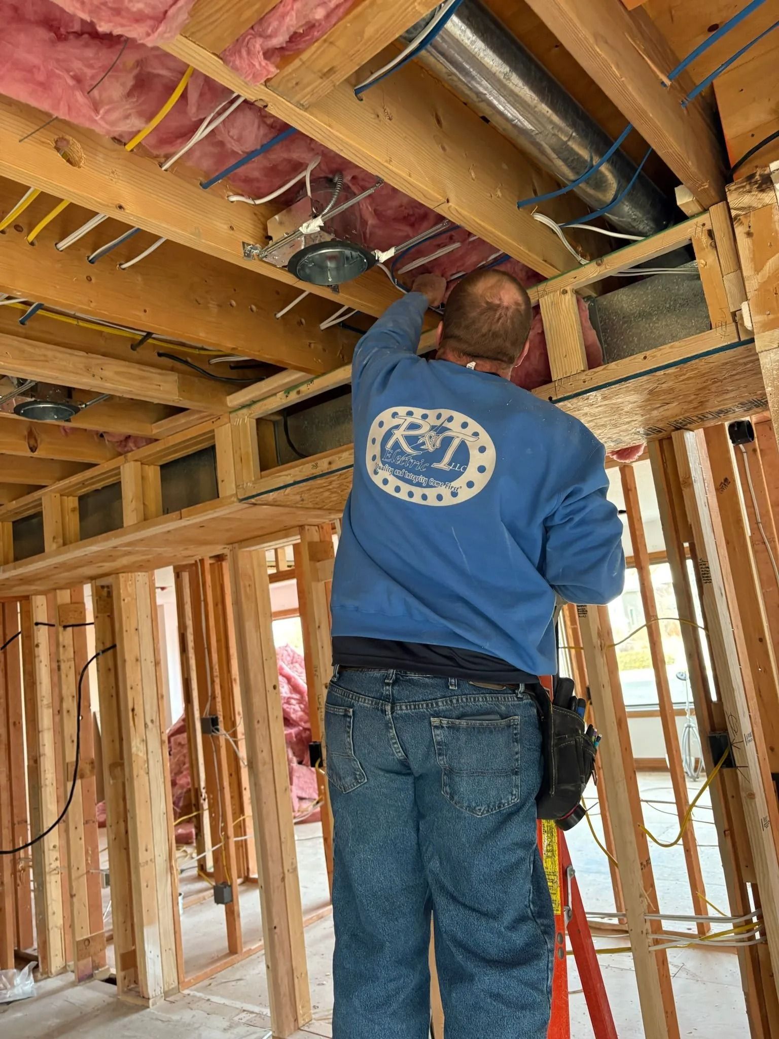 Man in blue sweatshirt working on electrical wiring in a construction site, standing on a ladder.