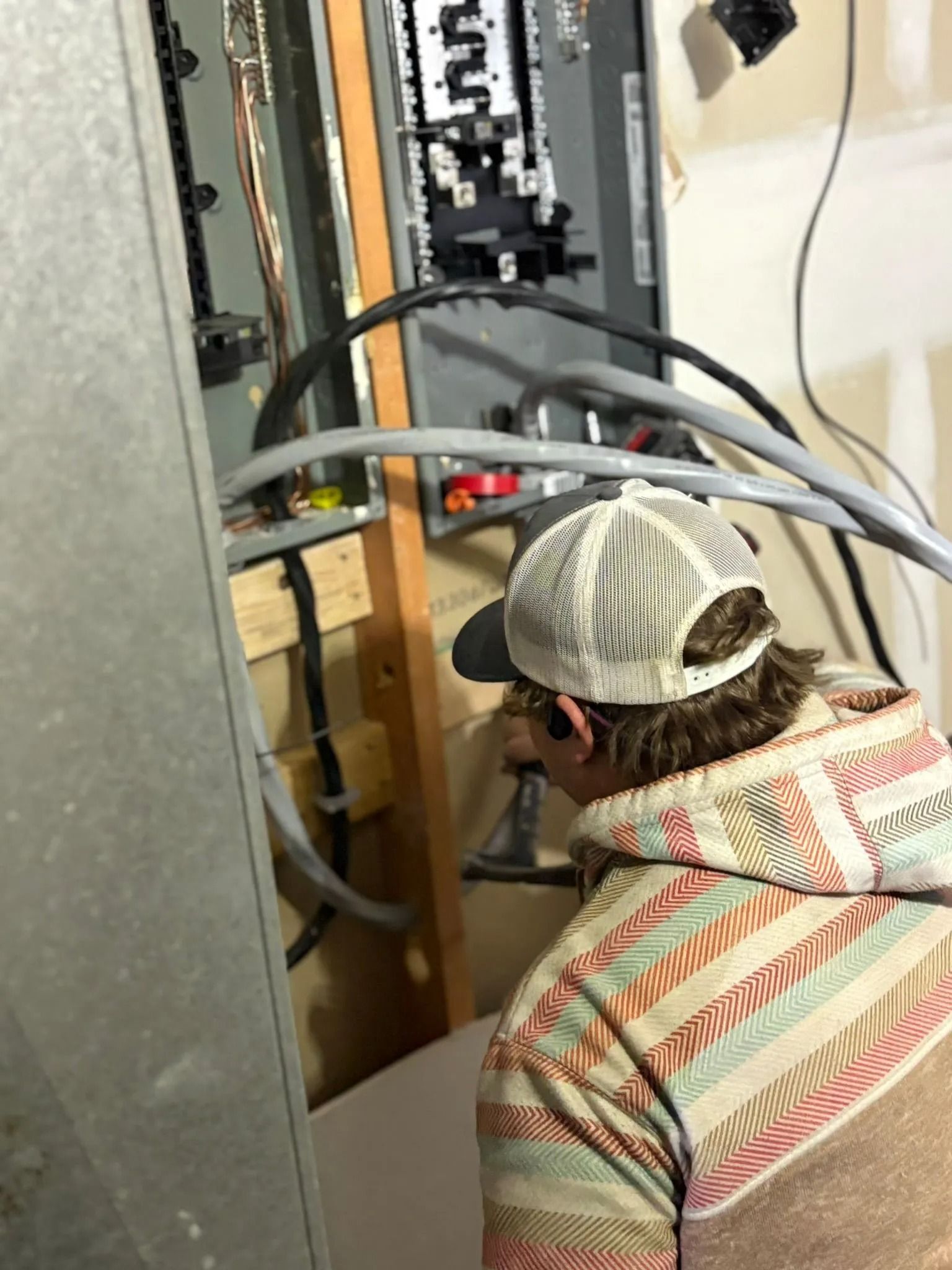 Electrician working on electrical panel, drilling into wood. Panel is gray, wires are visible.