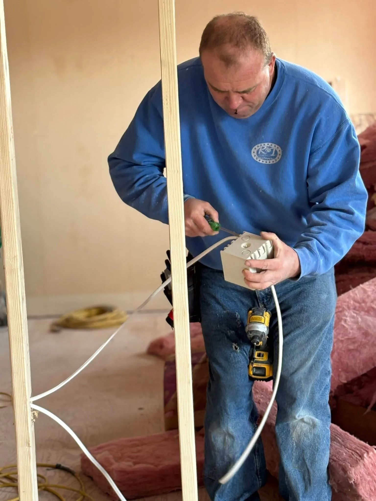 Man in blue sweatshirt and jeans wiring electrical box inside a room under construction.