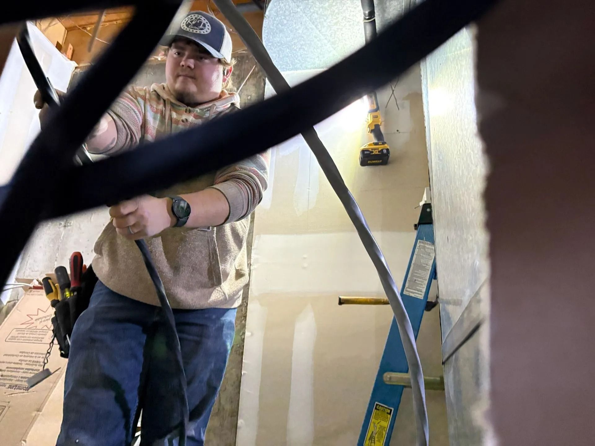 An electrician, in a construction site, pulls cable while standing on a ladder.