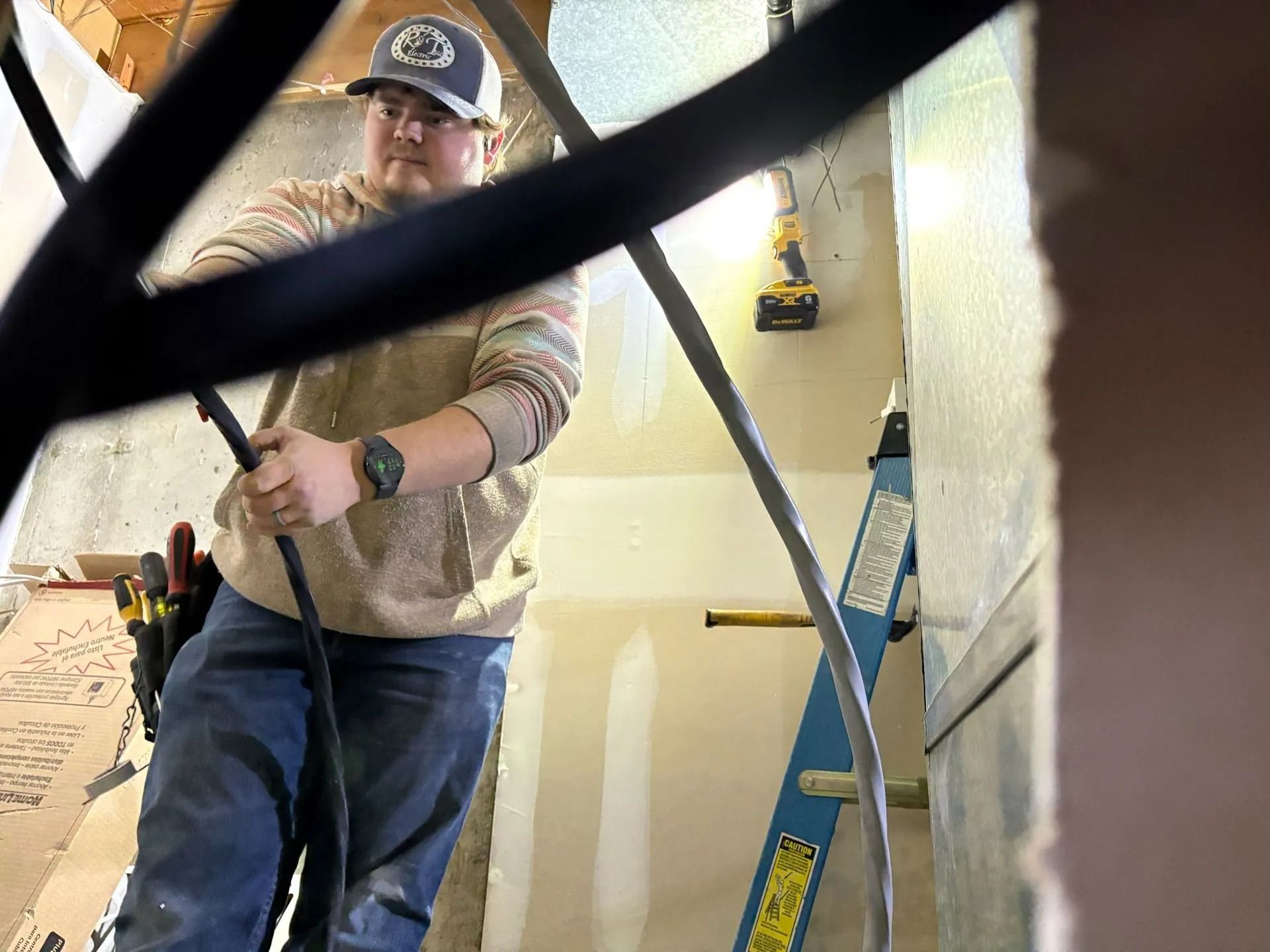 Electrician working on wiring, standing on a ladder. Wearing hat, hoodie, jeans, holding cable in a room.