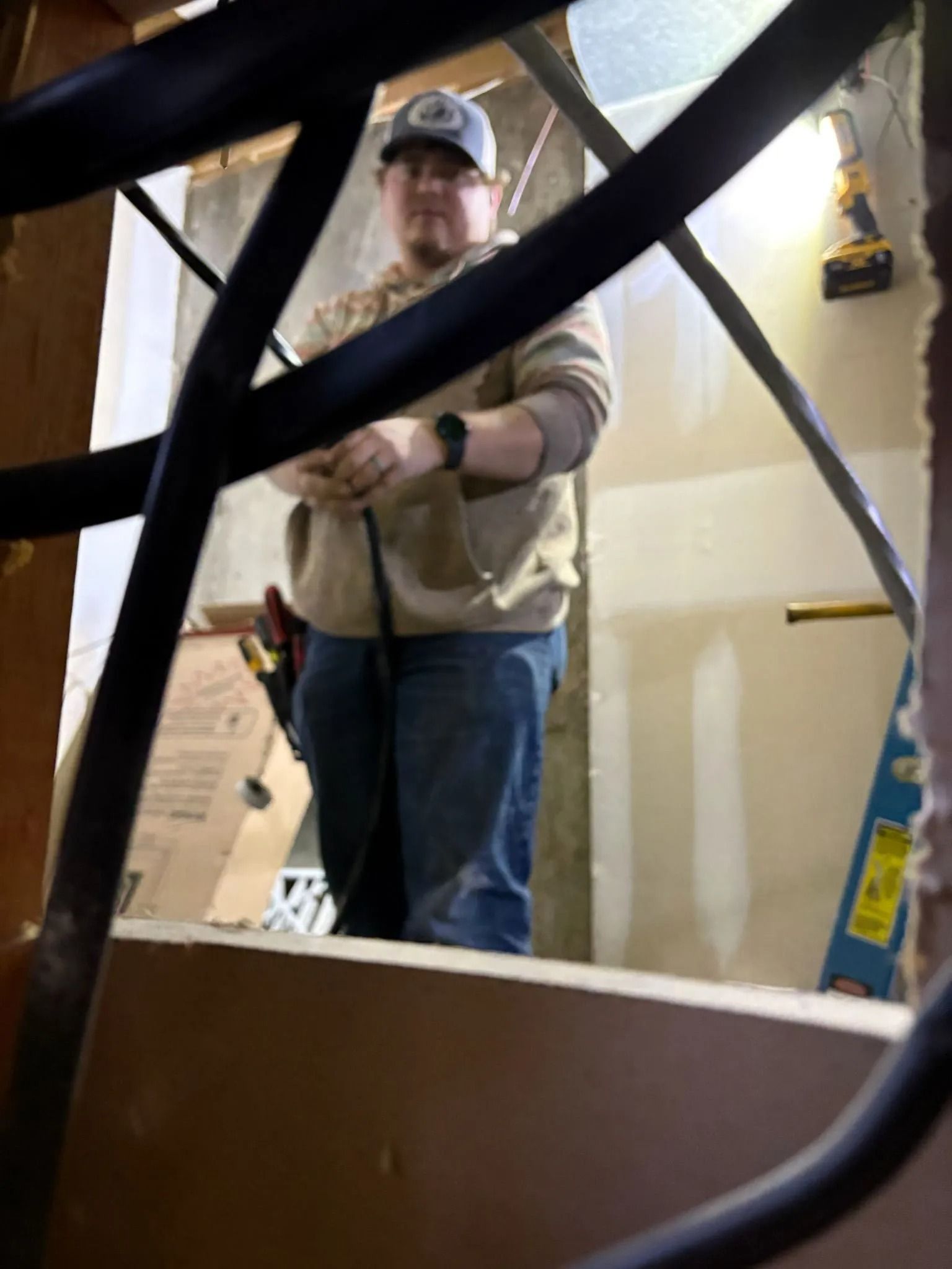 Man in tan hoodie and jeans working with wiring inside a wall, illuminated by a light.