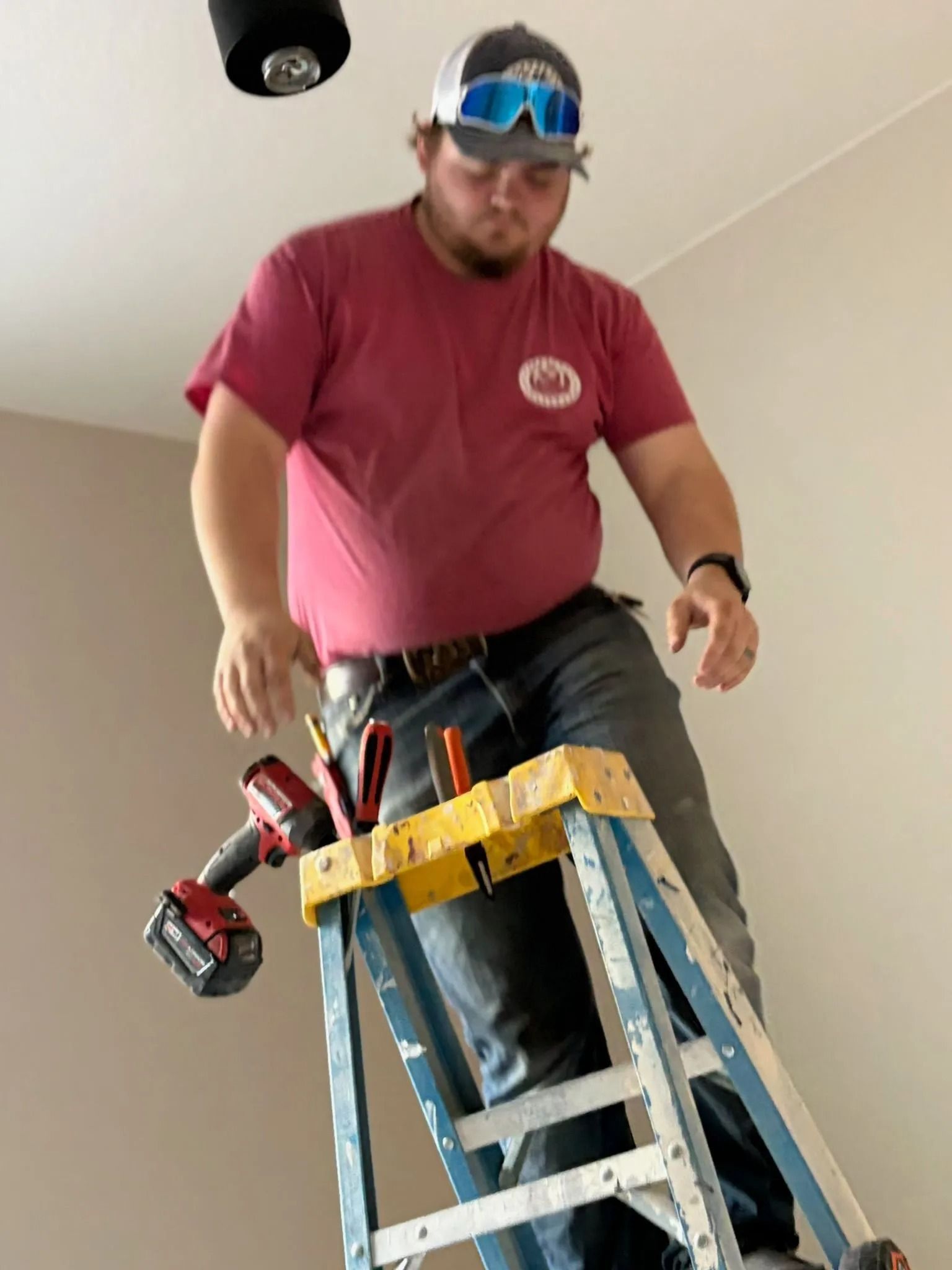 Man on stepladder, installing ceiling fixture. Tools on ladder, wearing hat and glasses, in a room.