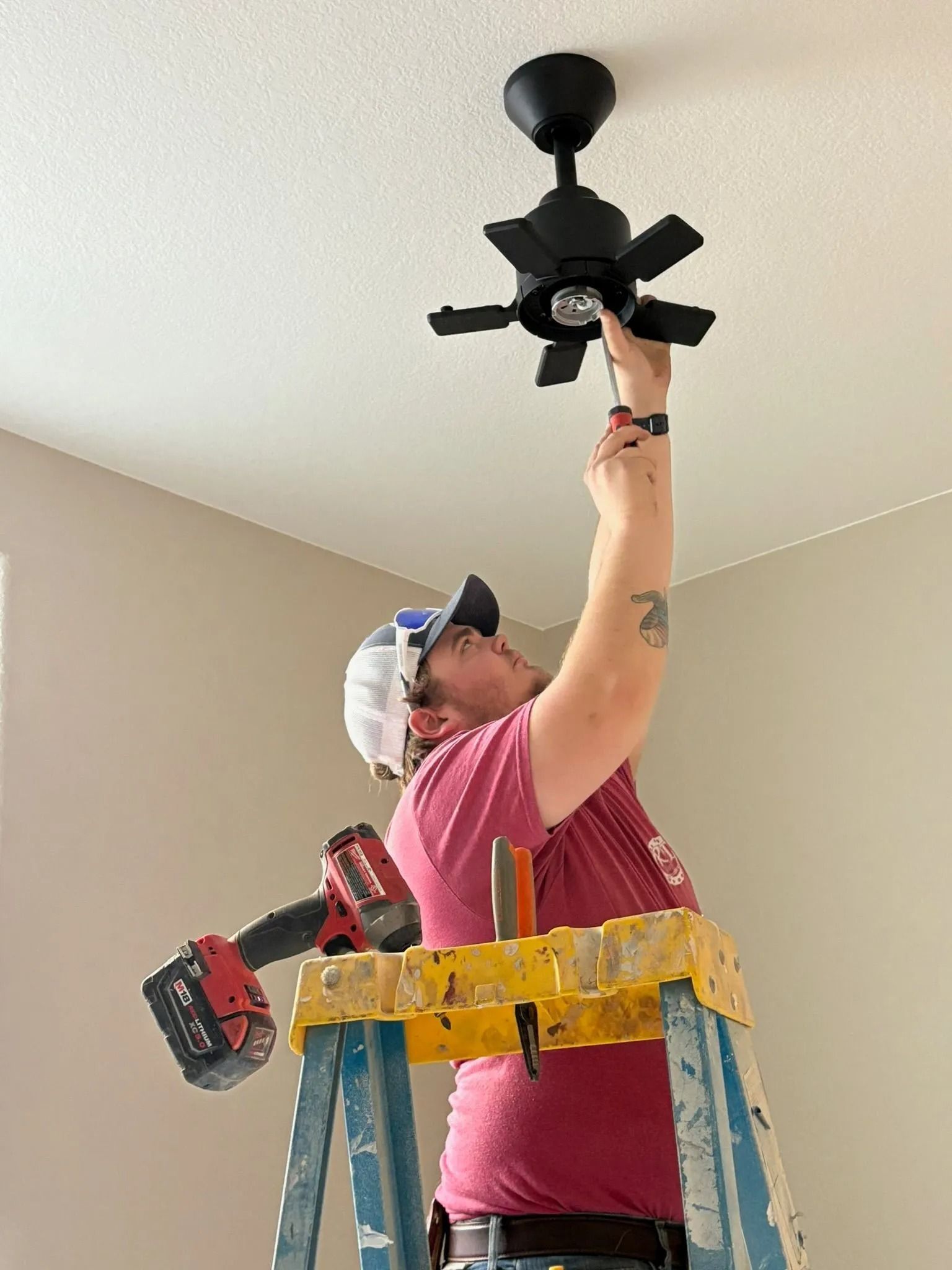 Person on a ladder installing a black ceiling fan, holding a tool. A red drill rests on the ladder.