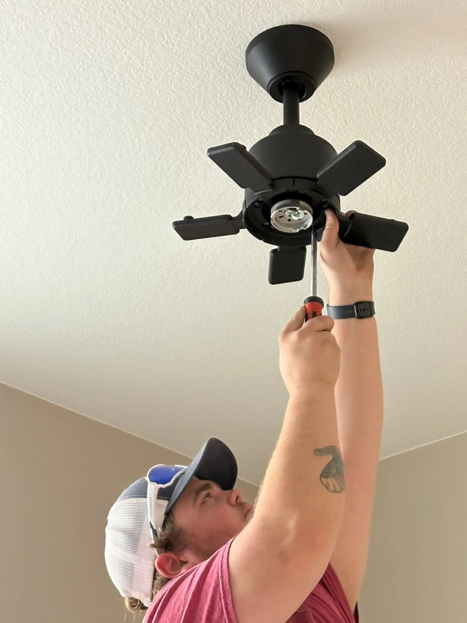 Person installing a black ceiling fan, using a screwdriver. Indoors, against a white ceiling.