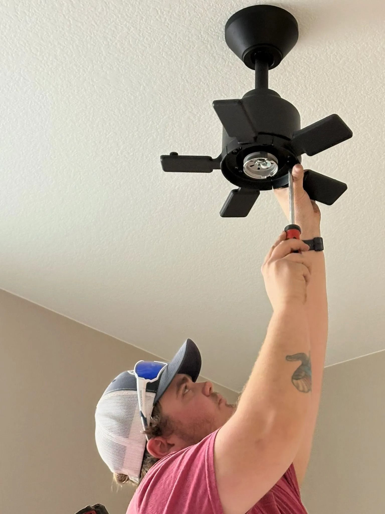 Person with hat installing a black ceiling fan, using a screwdriver. Indoors, against a white ceiling.