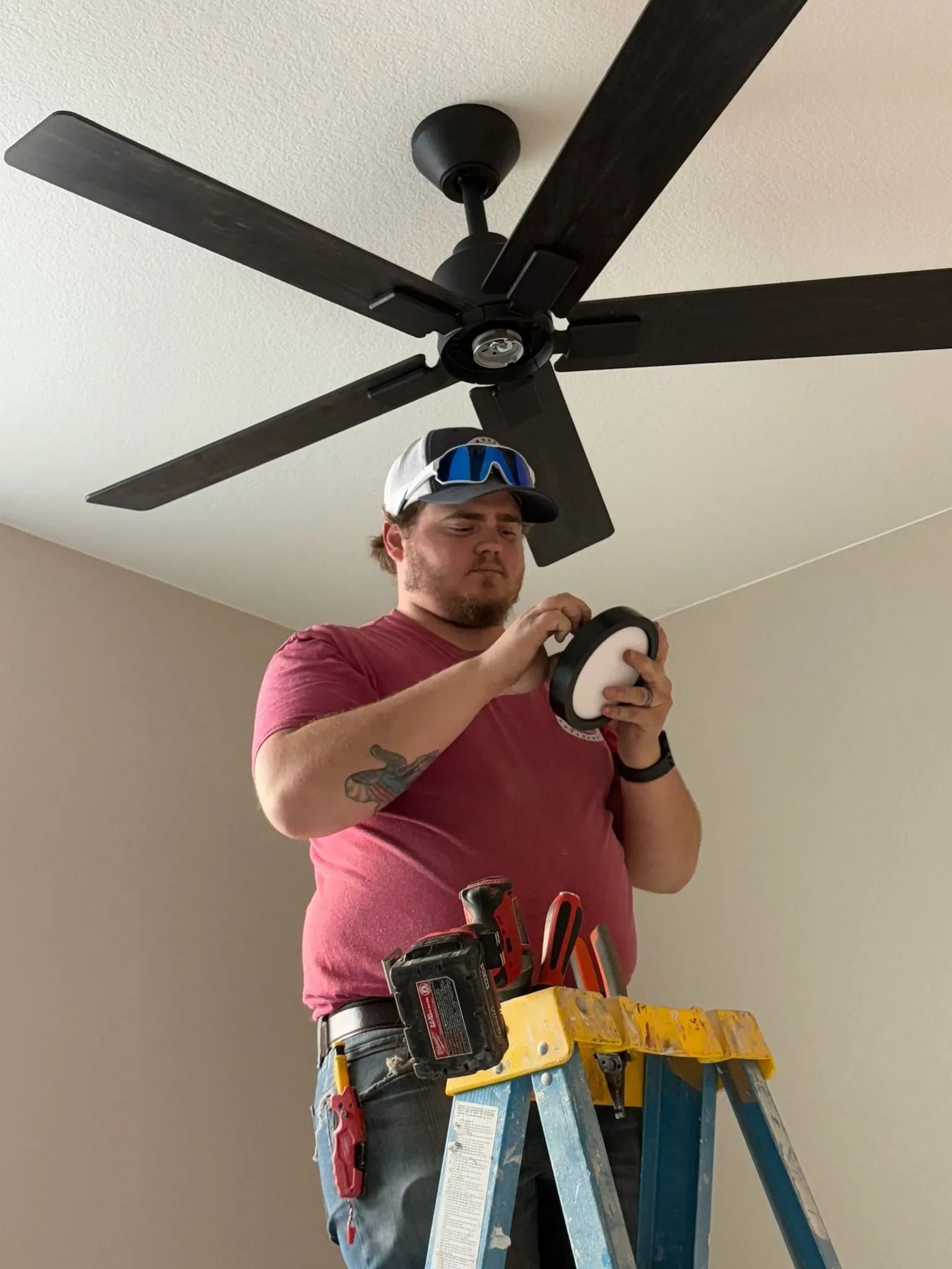 Man on a stepladder installing a ceiling fan light fixture. He is wearing a red shirt and a tool belt.