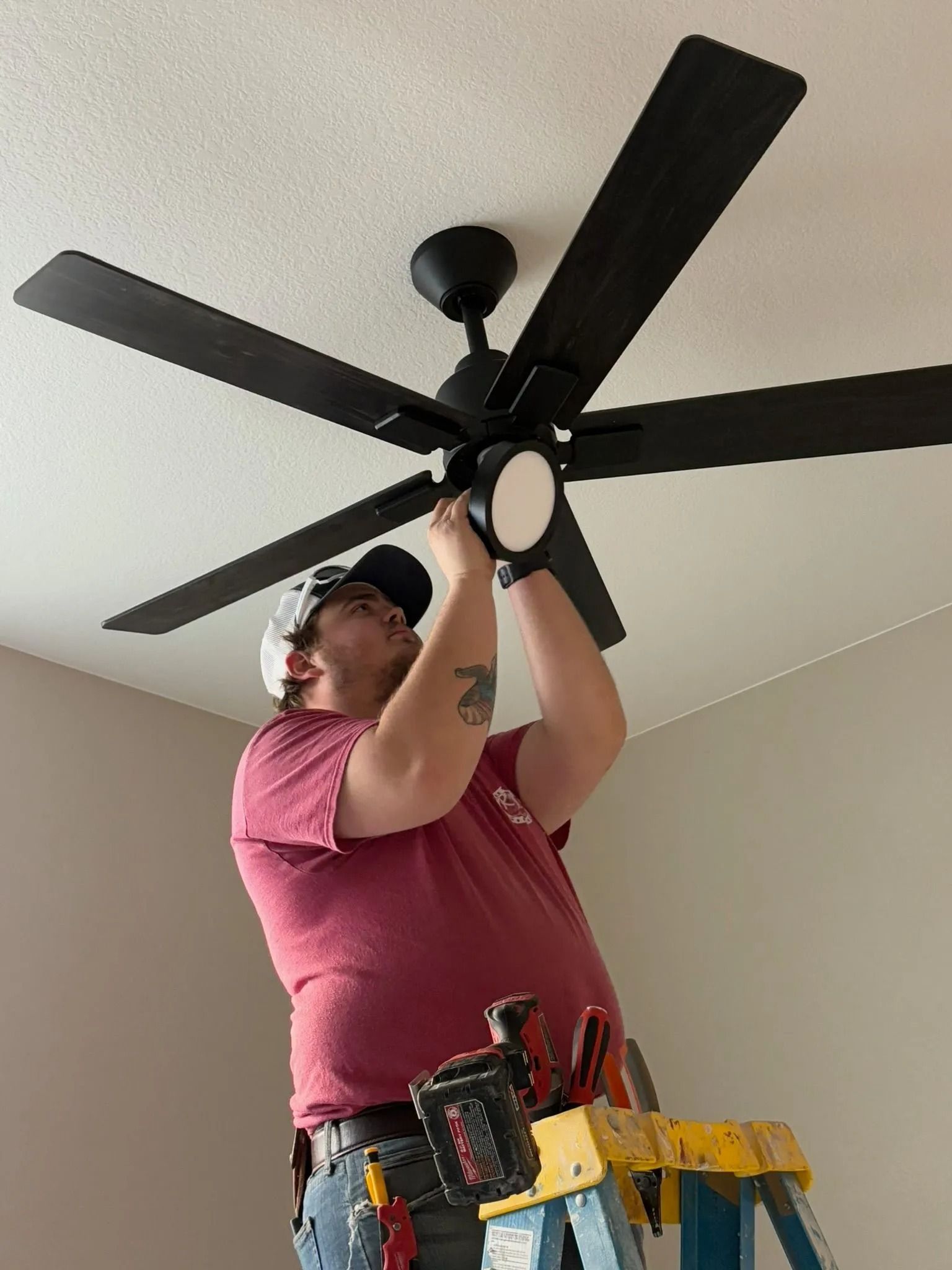 Man installing a black ceiling fan, standing on a ladder. Interior room, white ceiling, neutral walls.