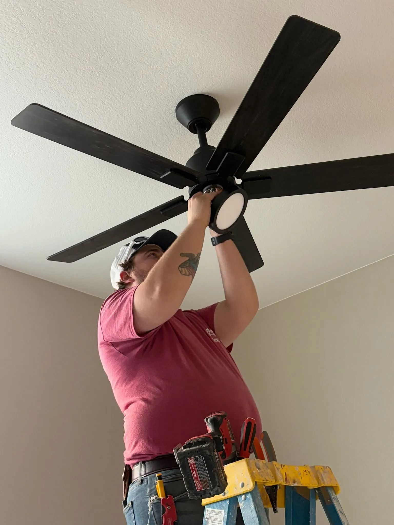 Person on a ladder installing a black ceiling fan with light in a room with white walls.