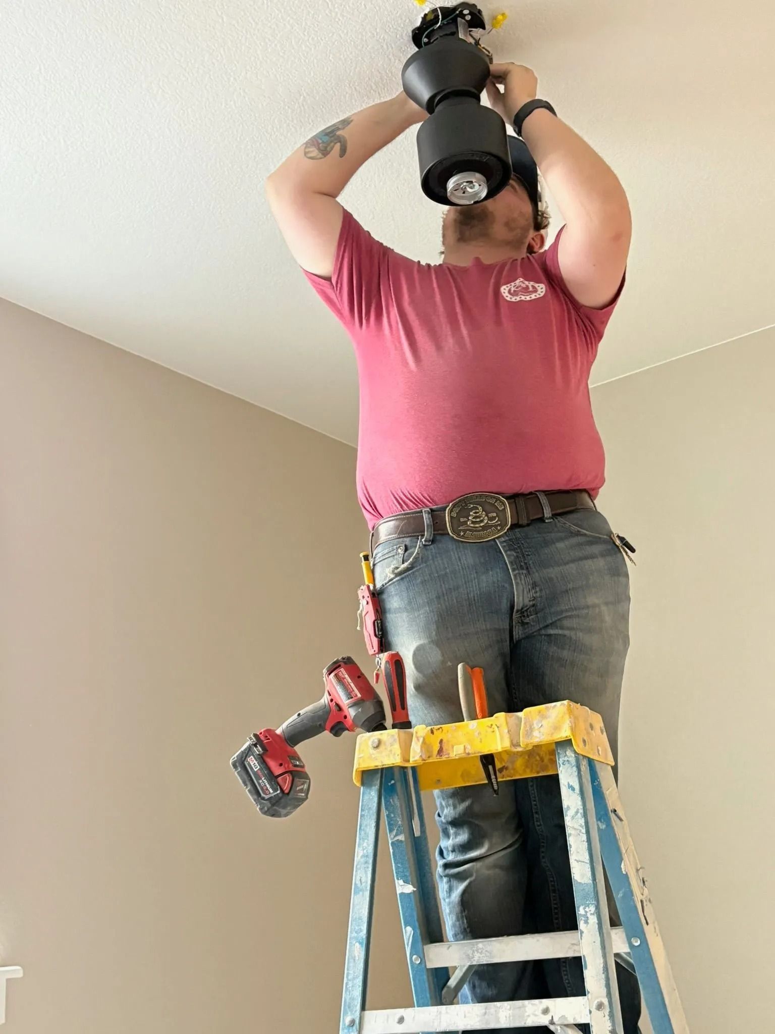 Person on ladder installing a ceiling fan. Wears red shirt, jeans. Tools on ladder. White ceiling, beige walls.