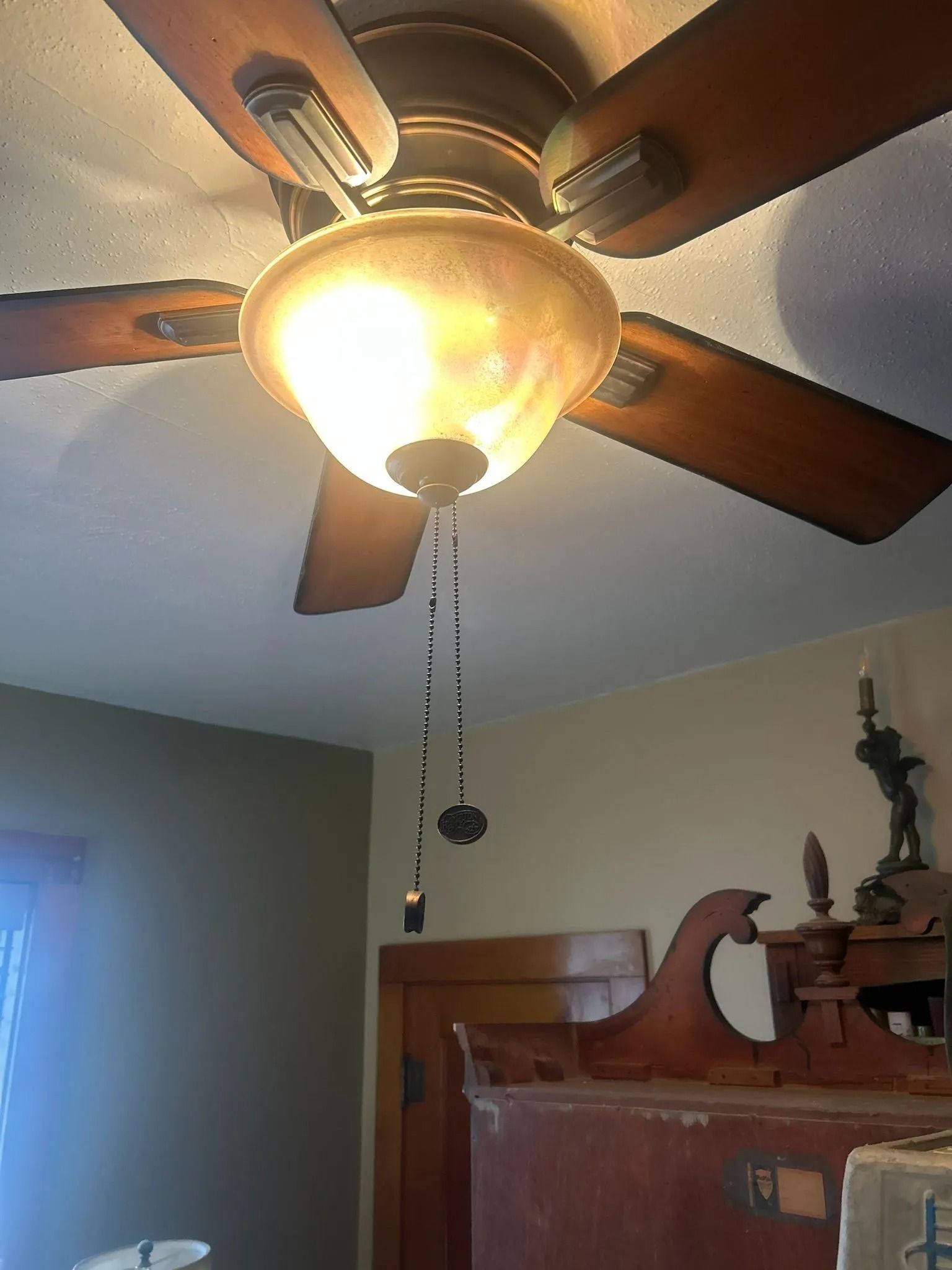 Ceiling fan with light fixture. Wooden blades, tan light, and pull chains hang down.