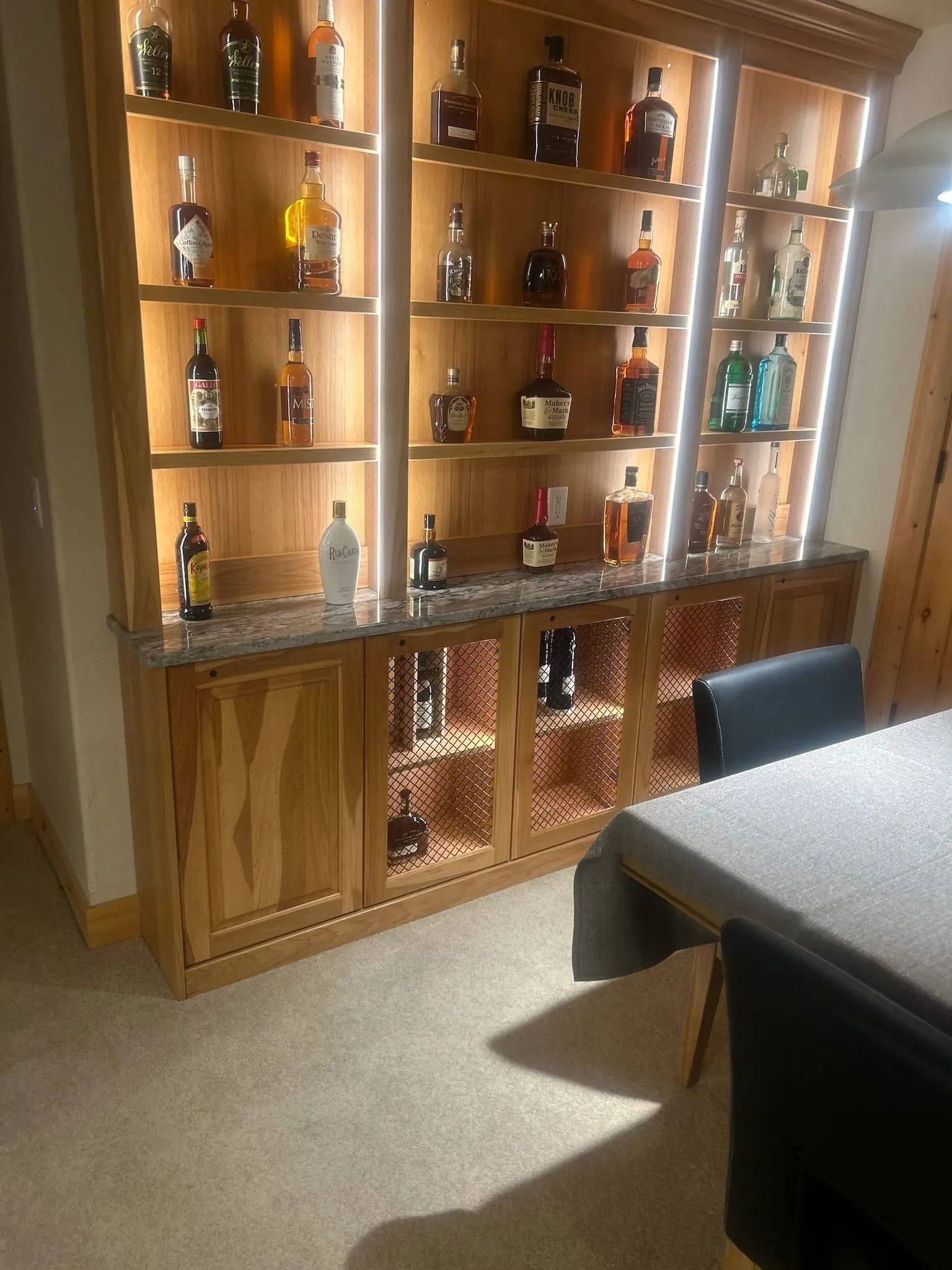 Wooden bar cabinet with liquor bottles lit by LED lights; stone countertop, lower cabinet with wire mesh.