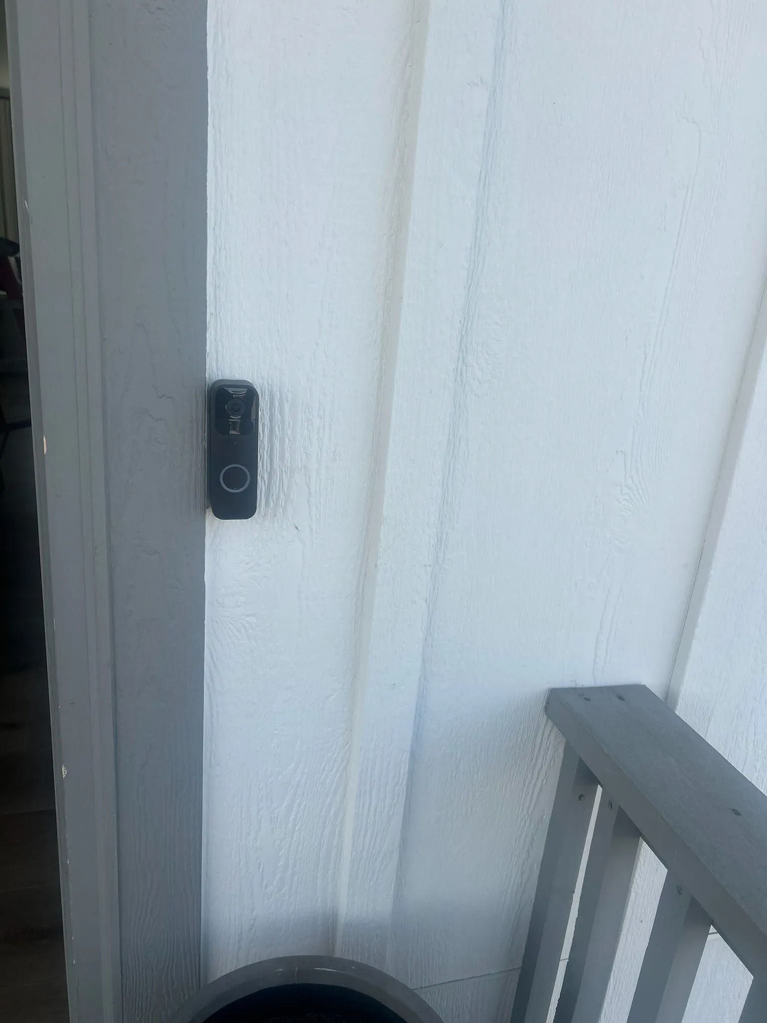 A black doorbell mounted on a white vertical siding wall next to a porch railing.