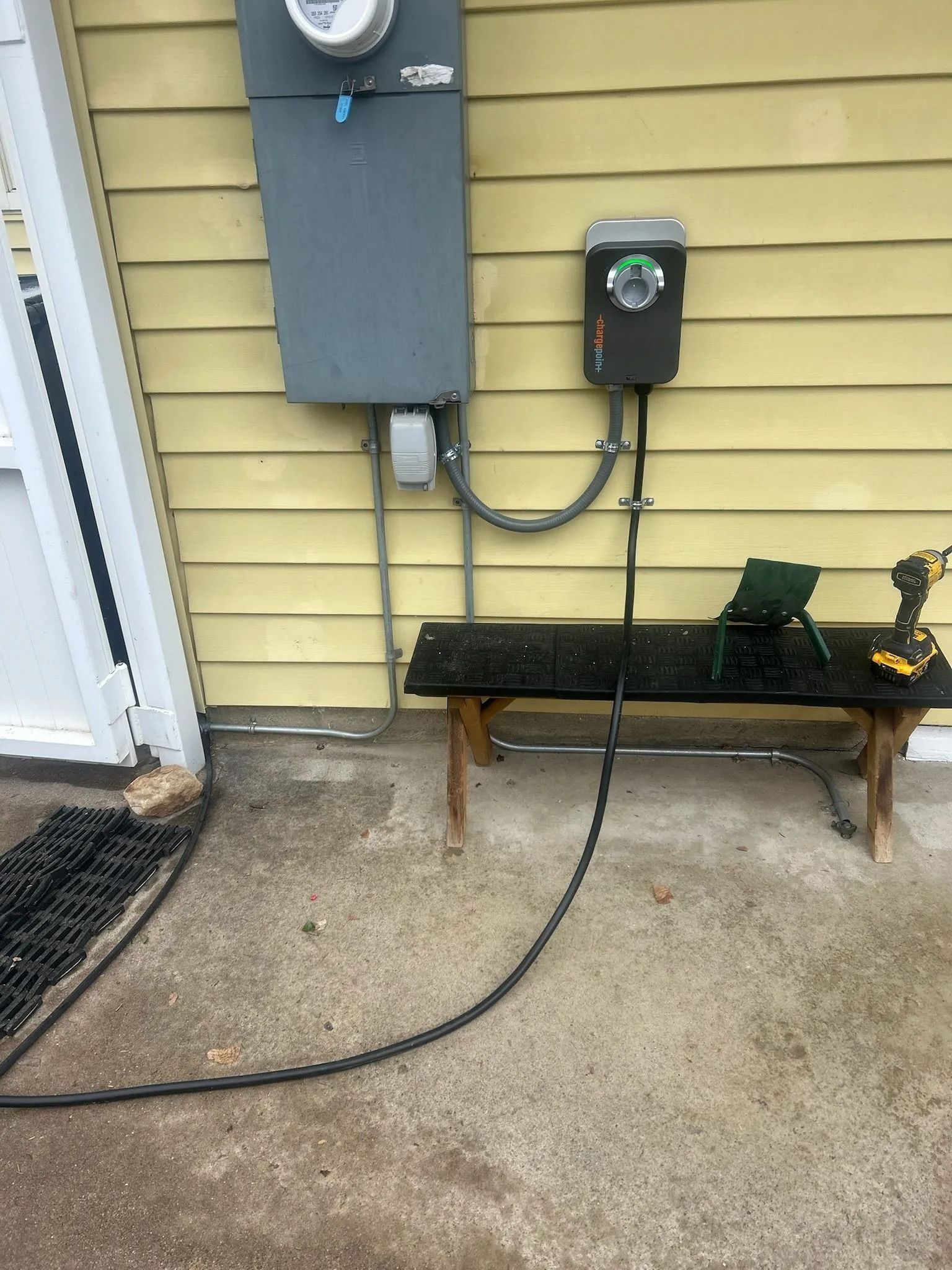 Exterior of a home with an electrical panel, charging station, and power cord running along the ground.