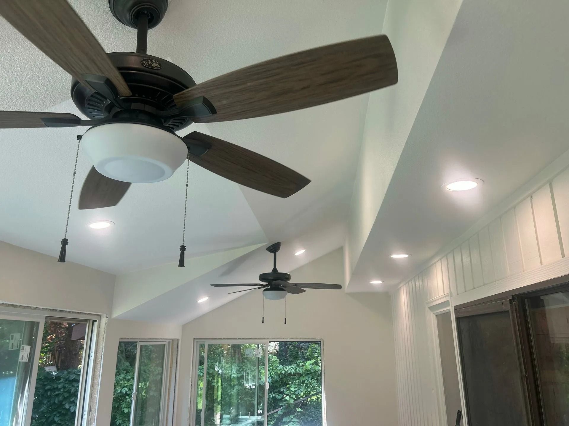 Two ceiling fans with lights in a room with recessed lighting and windows.
