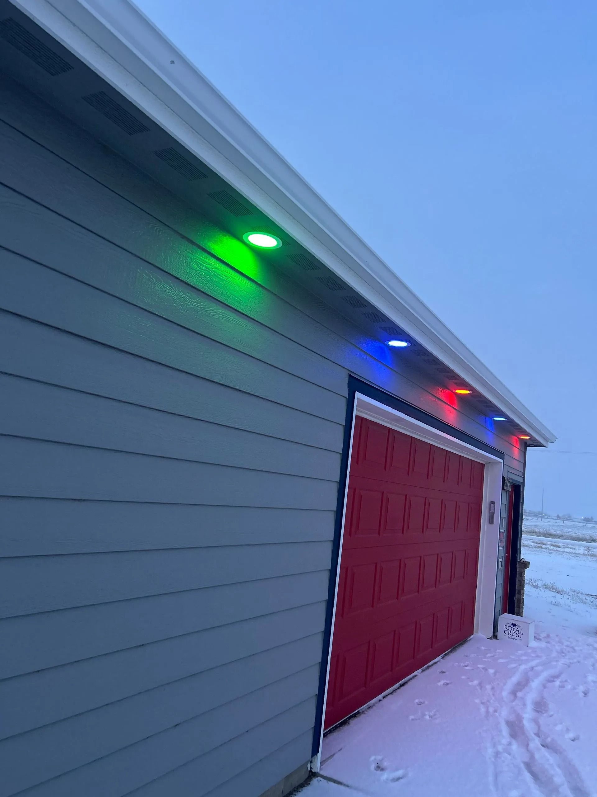 Garage with red door, blue-gray siding, and colorful lights along the roof in a snowy setting.