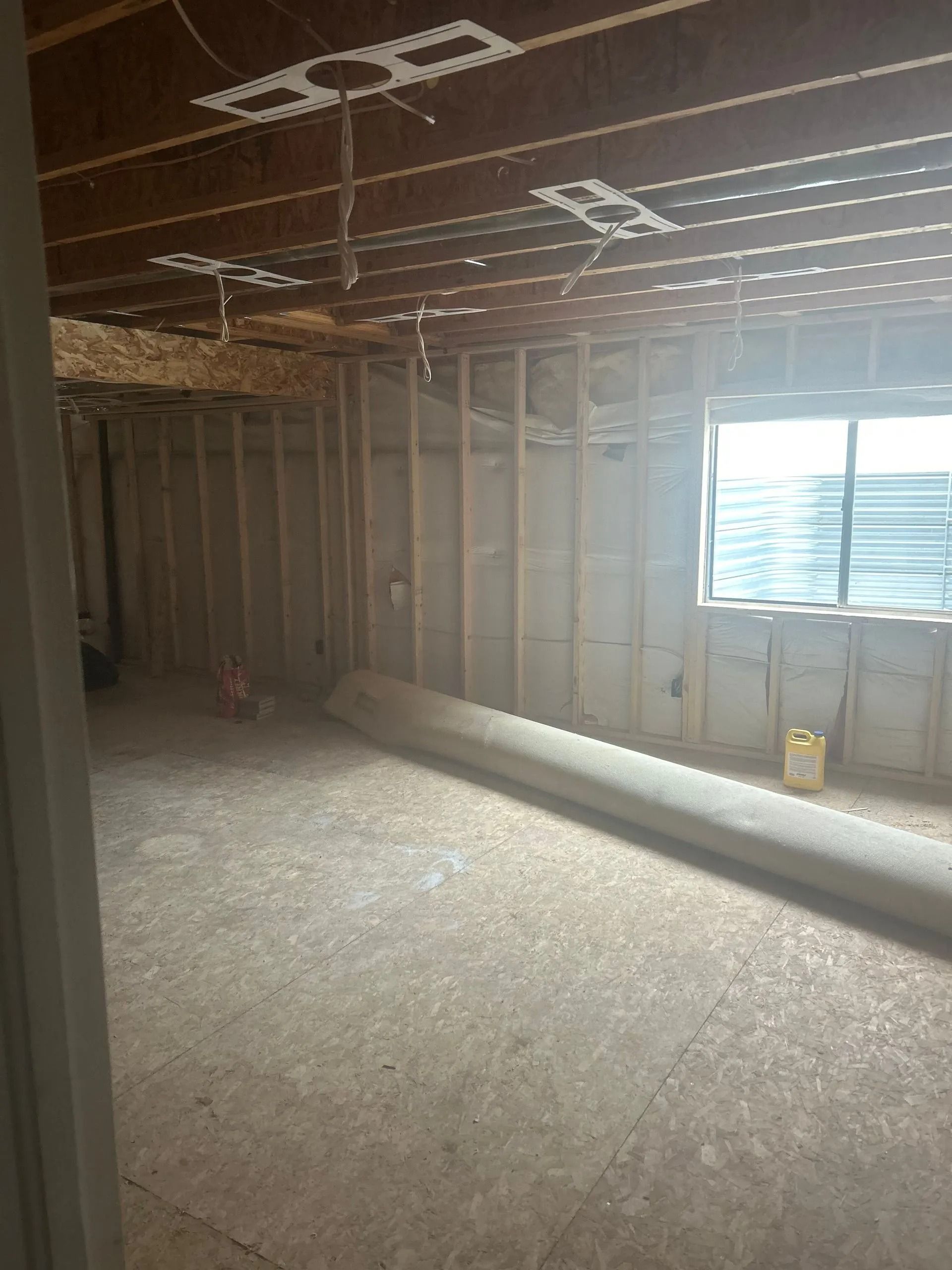Unfinished basement interior with exposed studs, concrete floor, and rolled-up carpet. Window on the right.